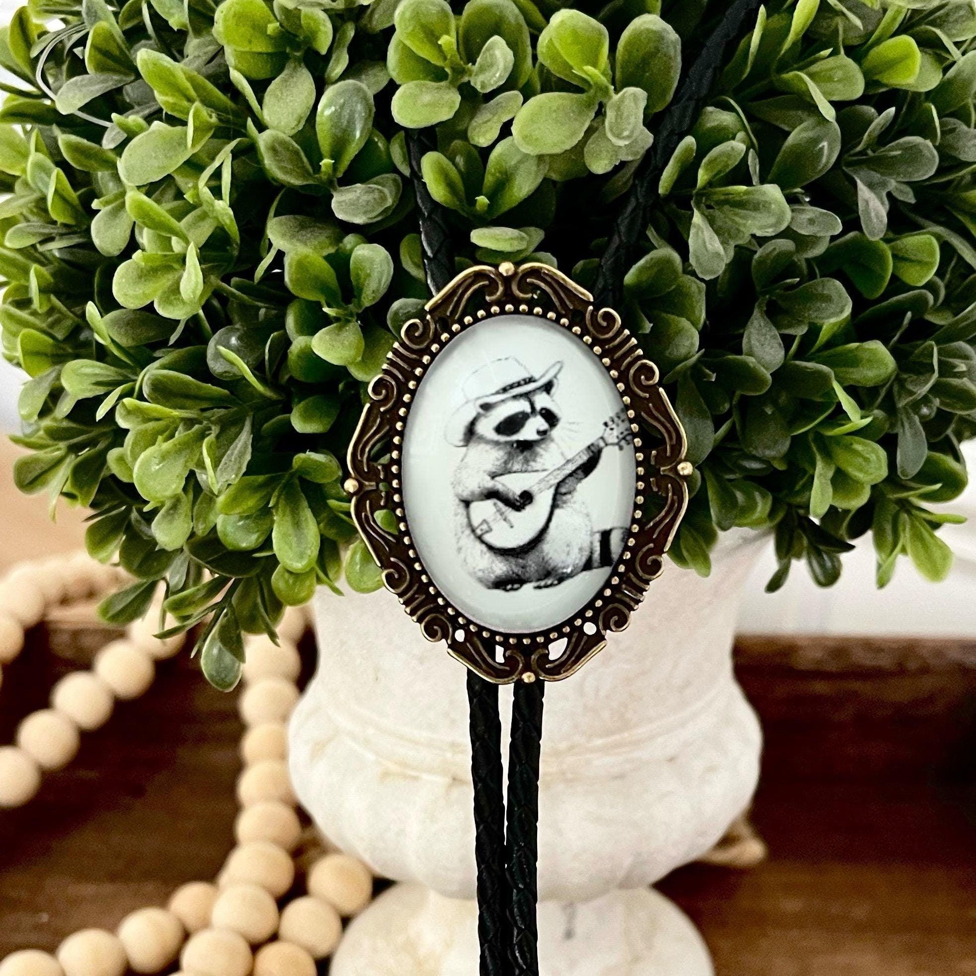 Raccoon bolo tie in black white and bronze hanging from a decorative green boxwood.