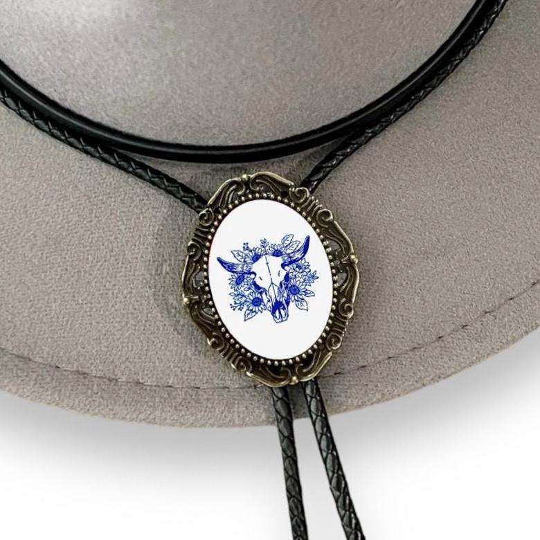 Infographic from The Bold Bow Tie titled Bolo Tie Features. Right: A vintage-style bolo tie with a blue and white cowboy riding a horse illustration inside an ornate bronze filigree frame on a black braided cord. Left: Lines connect the image to three checkmark features: Handmade, 39" Long Cord, and Pendant is 1.5" (L) x 1.25" (W). Bottom text reads Perfect Addition to Your Outfit.