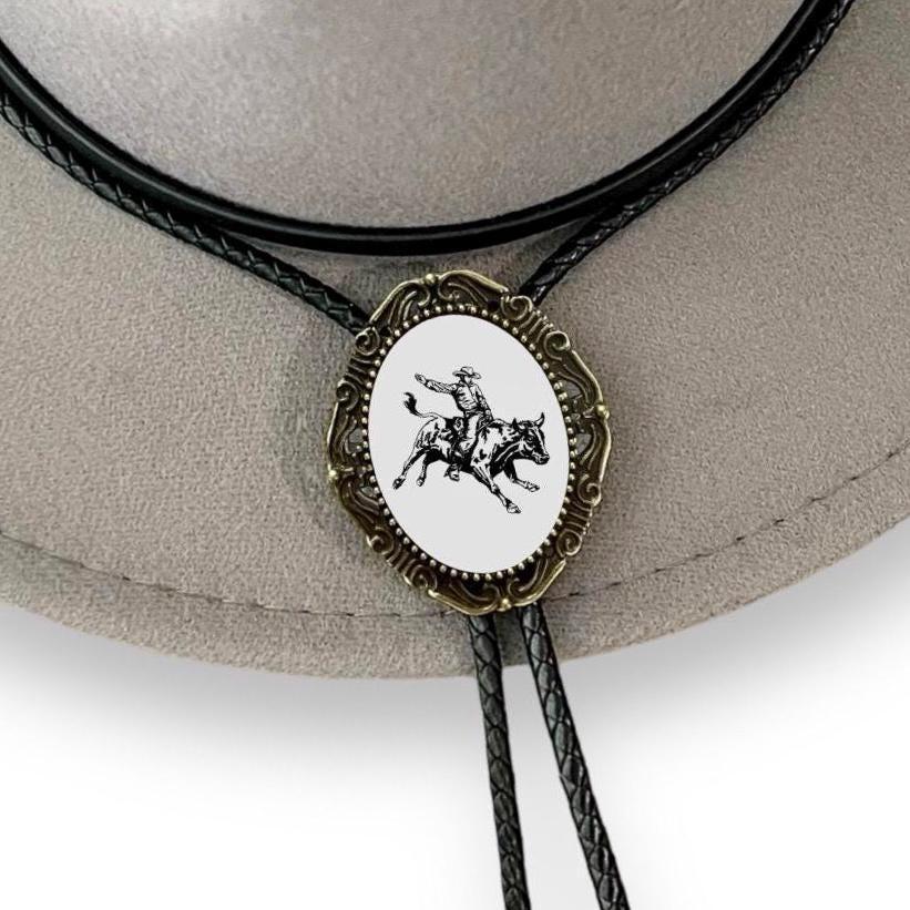 Infographic from The Bold Bow Tie titled Bolo Tie Features. Right: A vintage-style bolo tie with a blue and white cowboy riding a horse illustration inside an ornate bronze filigree frame on a black braided cord. Left: Lines connect the image to three checkmark features: Handmade, 39" Long Cord, and Pendant is 1.5" (L) x 1.25" (W). Bottom text reads Perfect Addition to Your Outfit.