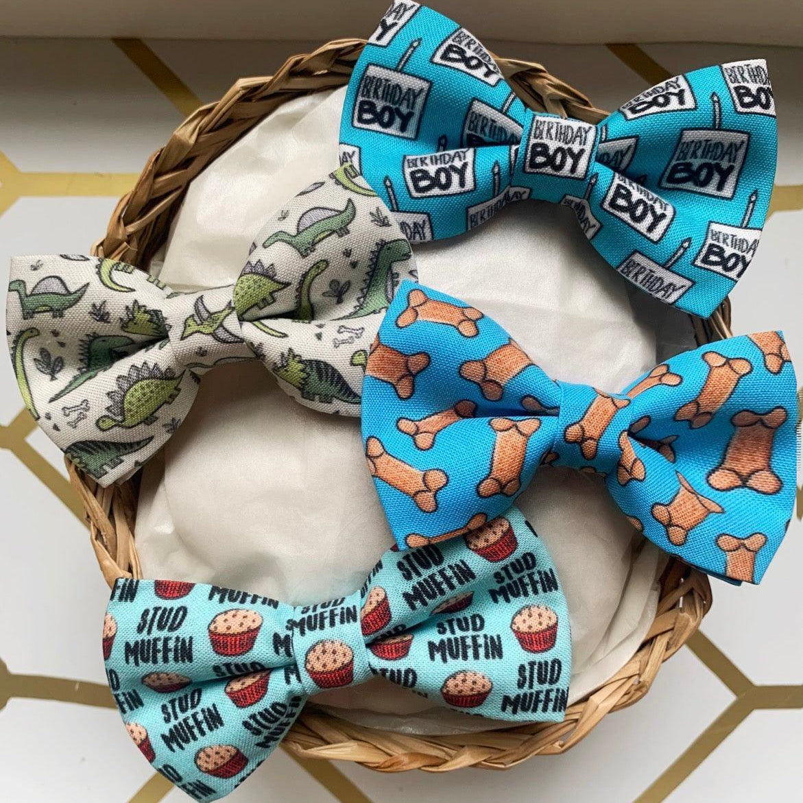 Gift set of three pet bow ties in blue birthday boy print pale dinosaur print and muffin print arranged in a wicker basket