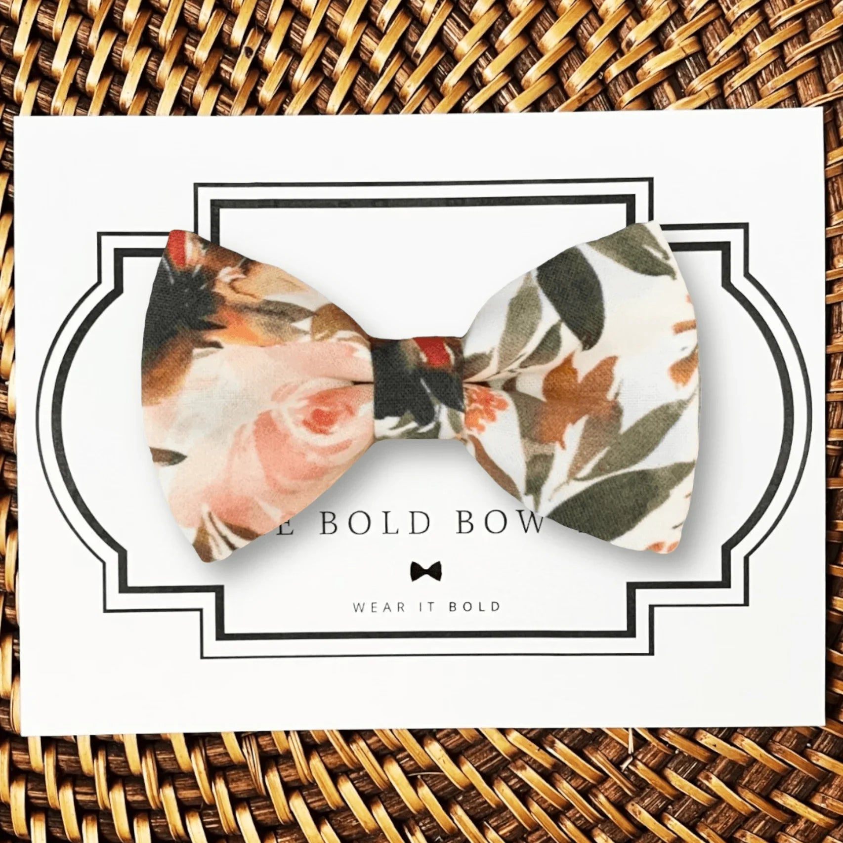 A dog bow tie with a floral pattern in terracotta and ivory colors, displayed on a card with 'THE BOLD BOW TIE' branding.