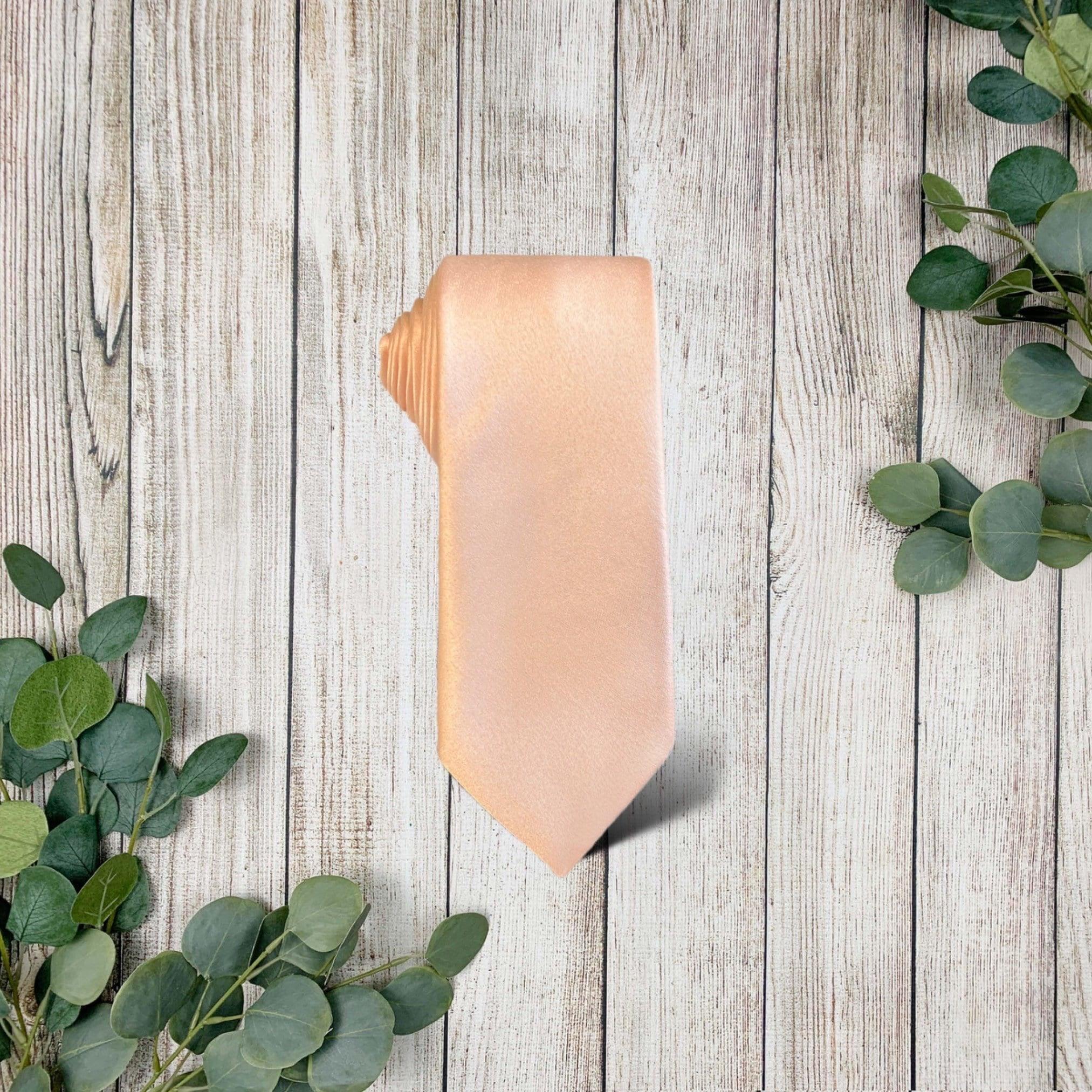 A blush pink tie for men laid out on a wooden surface surrounded by green foliage.
