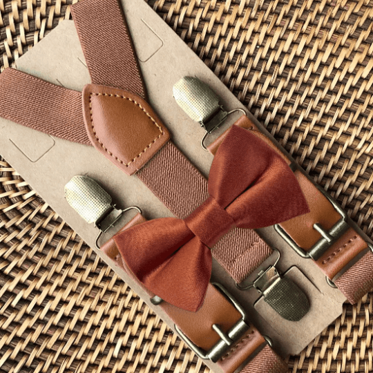 A set of terracotta bow tie and cognac brown faux leather suspenders displayed on a textured surface.