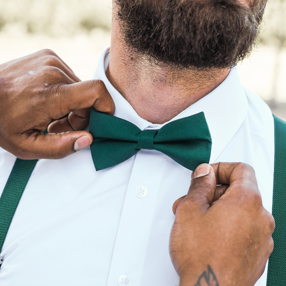 Groomsman wearing dark green suspenders and putting on a juniper green bow tie for a wedding.