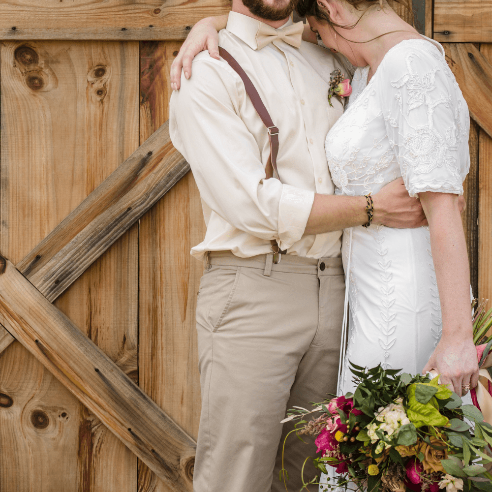 Groom wearing champagne bow tie at his wedding in front of a barn door, lovingly laughing with his bride.