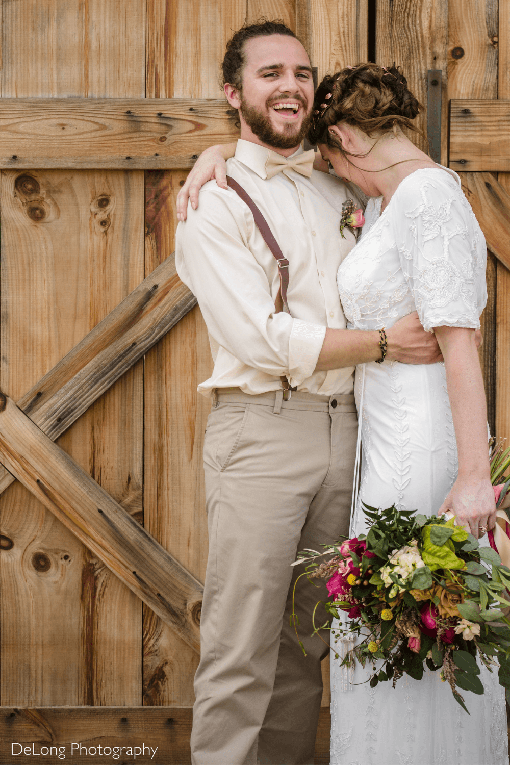 Groom wearing champagne bow tie at his wedding in front of a barn door, lovingly laughing with his bride.