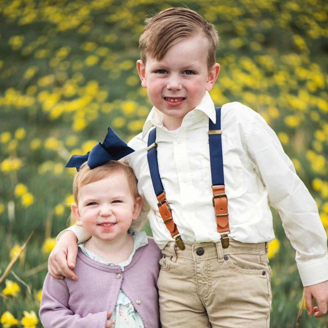 Ring bearer navy suspenders for wedding worn by a toddler and hugging the flower girl at a wedding.