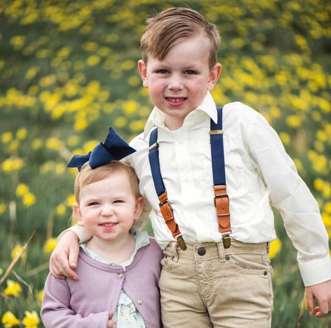 Ring bearer navy suspenders for wedding worn by a toddler and hugging the flower girl at a wedding.