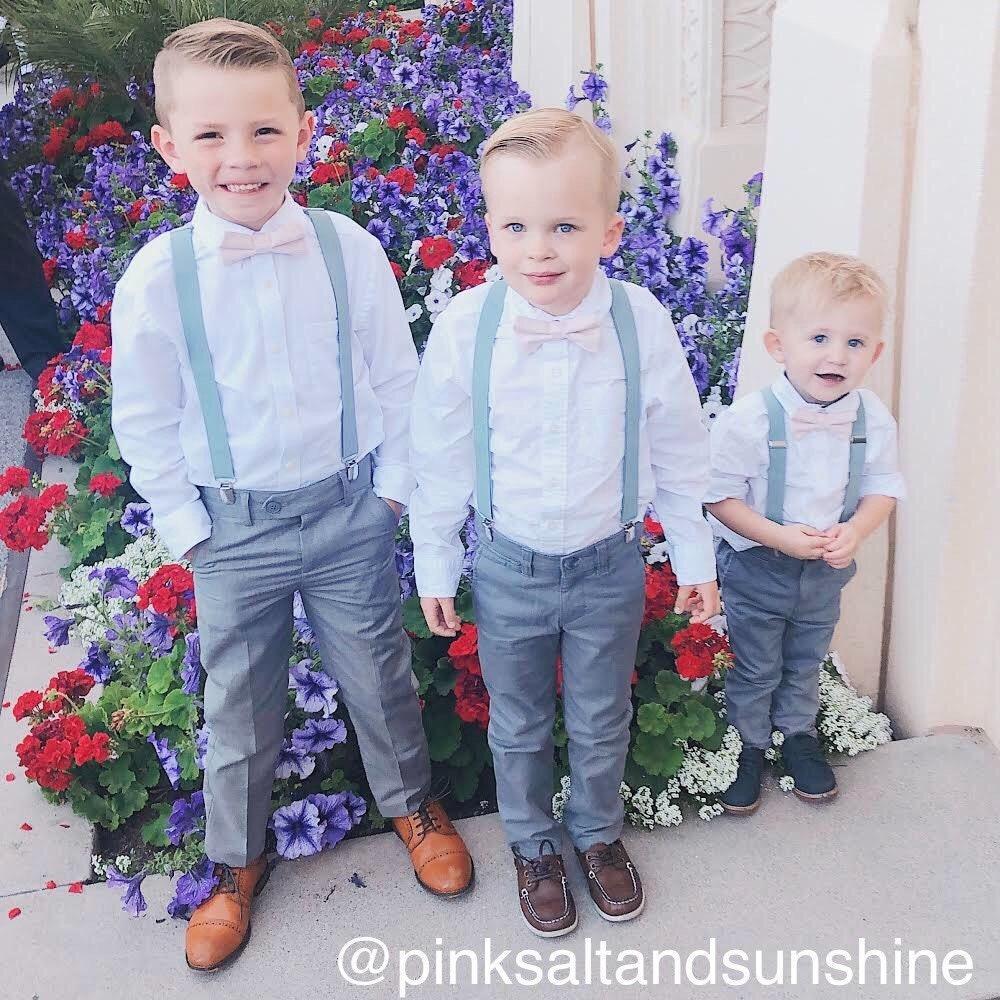 Three boys in light blue shirts blush bow ties and gray suspenders standing among colorful flowers outside