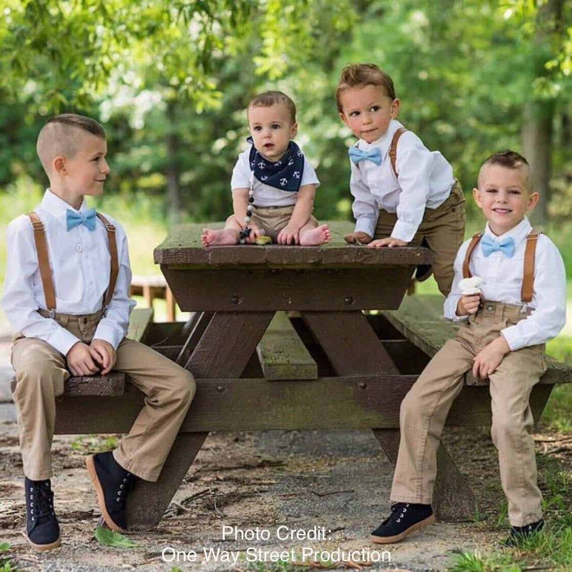 Four young boys wear white shirts with cornflower blue bow ties and brown leather suspenders, posing on a wooden picnic table outdoors.