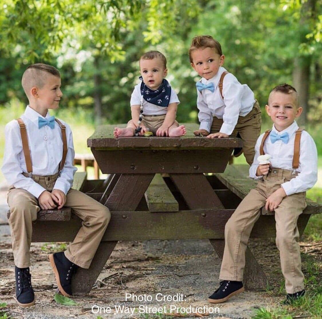 Four young boys wear white shirts with cornflower blue bow ties and brown leather suspenders, posing on a wooden picnic table outdoors.