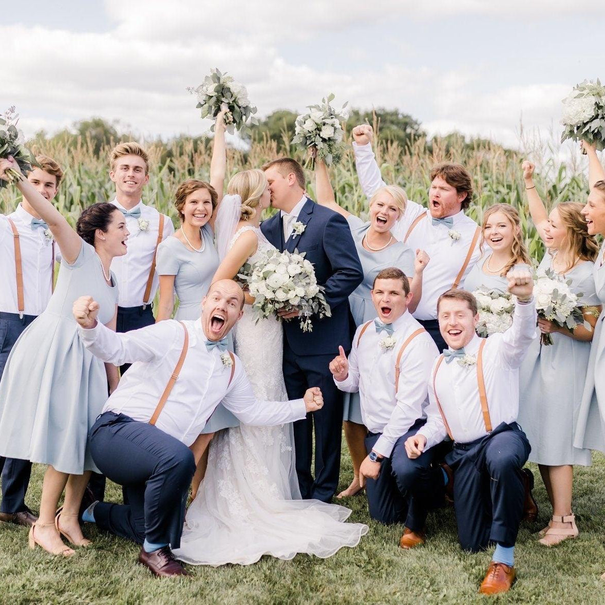 Group wedding party posing in a field among corn stalks wearing pale blue dresses and tan suspenders with matching slate bow ties