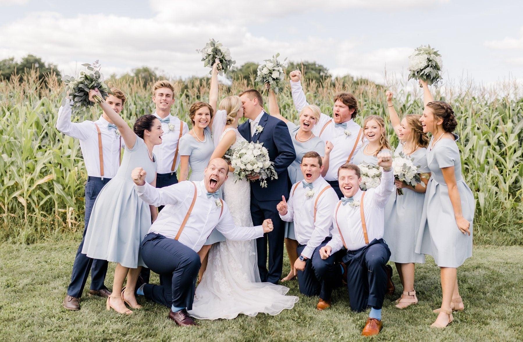 Group wedding party posing in a field among corn stalks wearing pale blue dresses and tan suspenders with matching slate bow ties