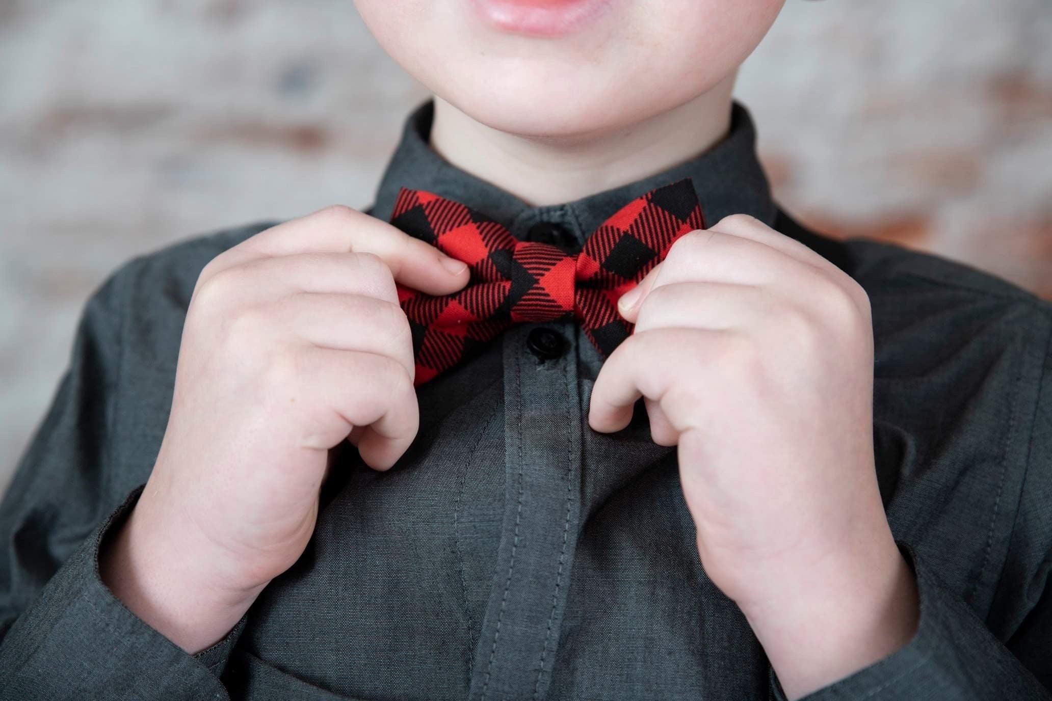 Close up of a child adjusting a red and black buffalo plaid bow tie against a dark shirt.