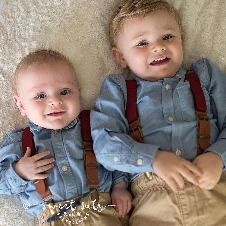 Two toddlers wearing burgundy suspenders with brass clips over light blue shirts, lying on a soft beige surface.