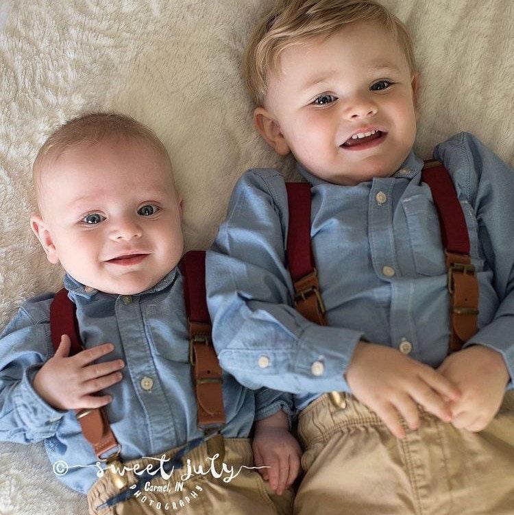 Two toddlers wearing burgundy suspenders with brass clips over light blue shirts, lying on a soft beige surface.