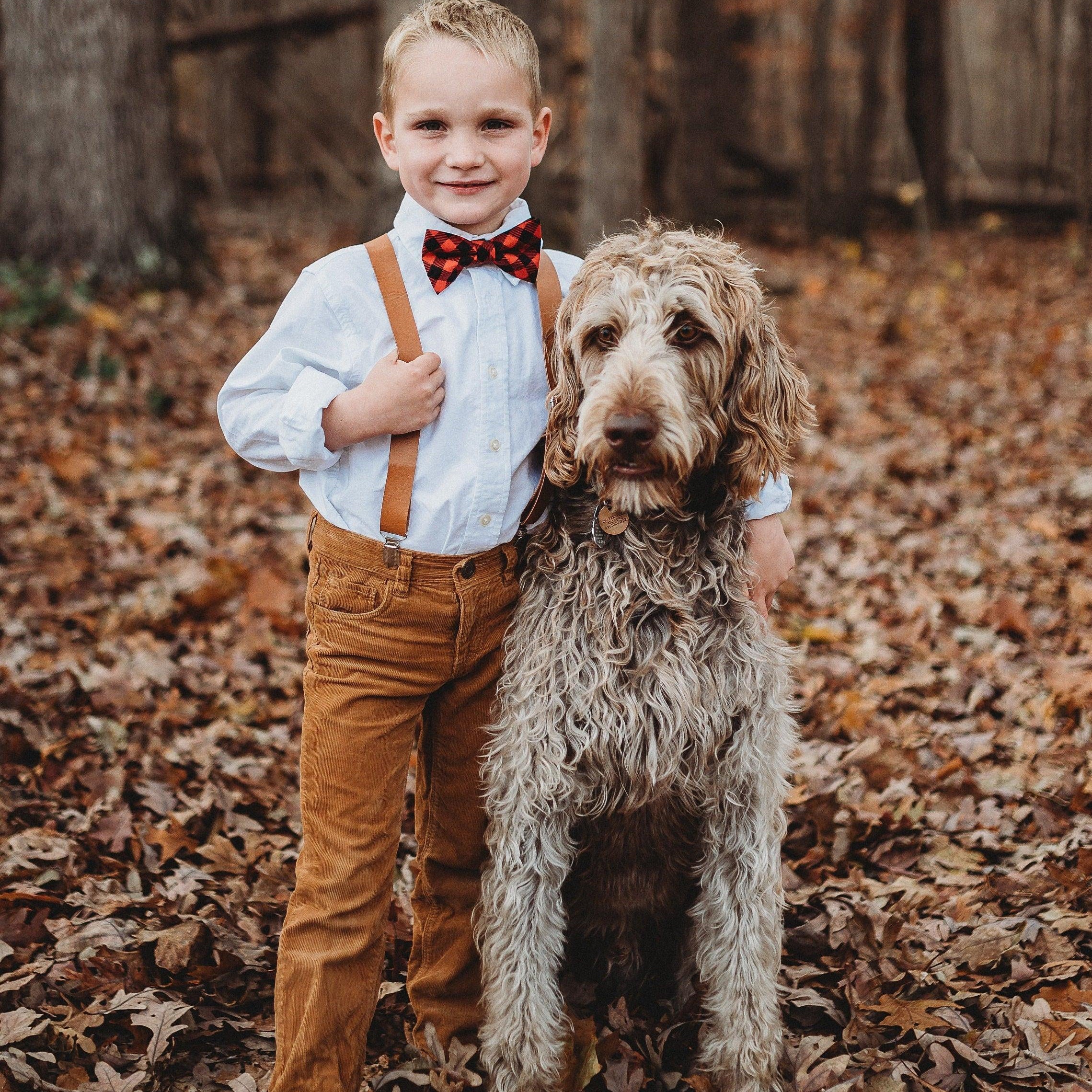 Child wearing buffalo plaid bow tie with brown vegan leather suspenders outdoors among fallen leaves with a dog.