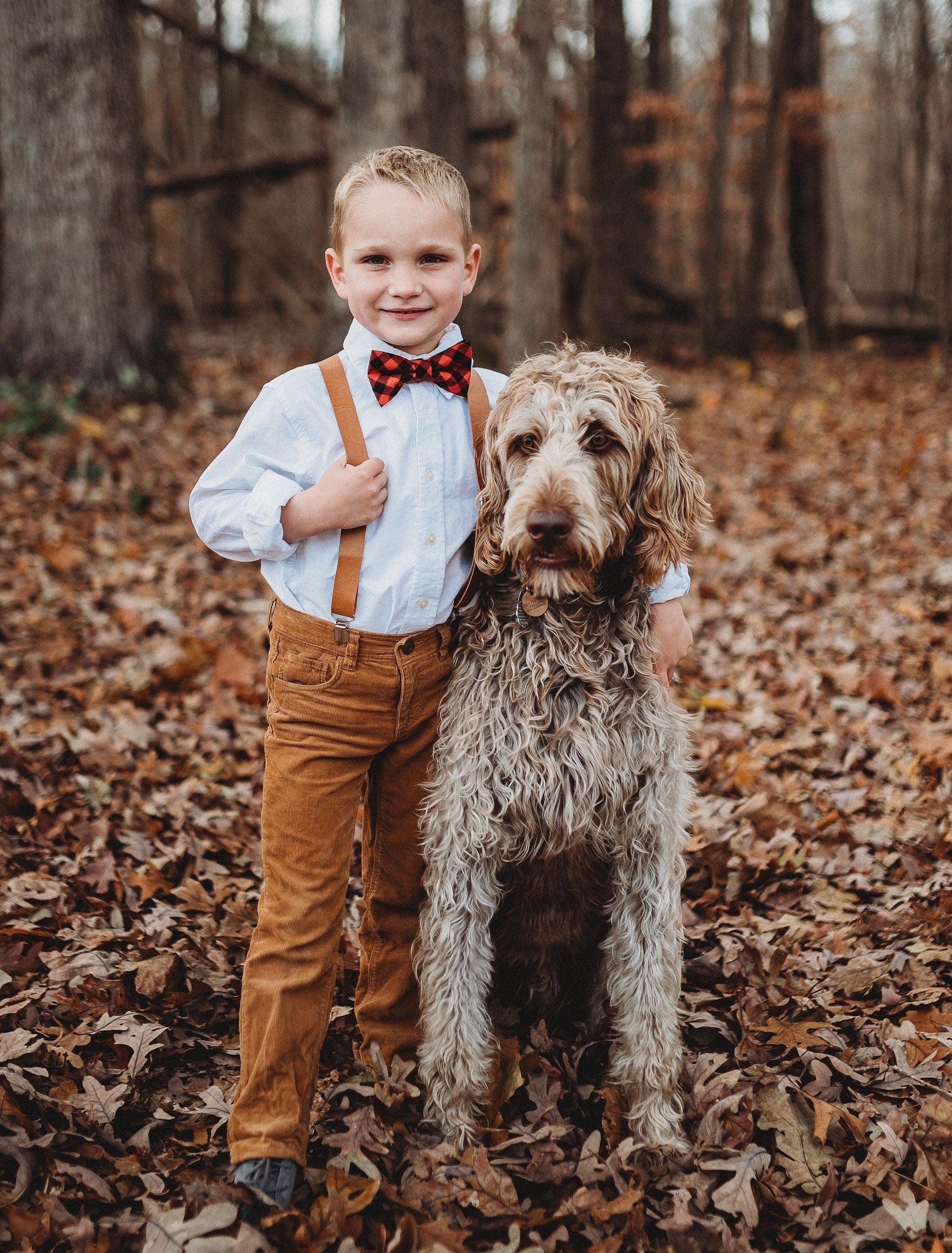 Child wearing buffalo plaid bow tie with brown vegan leather suspenders outdoors among fallen leaves with a dog.
