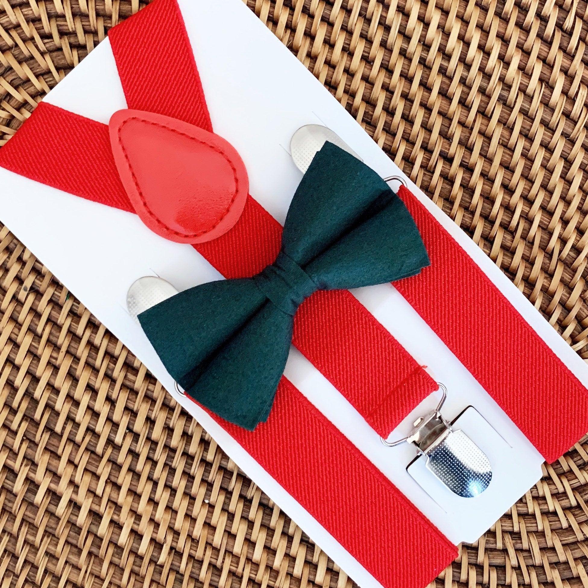 Close up of red suspenders and red bow tie on a white display card, with a dark green bow tie and metal clips on a woven mat.