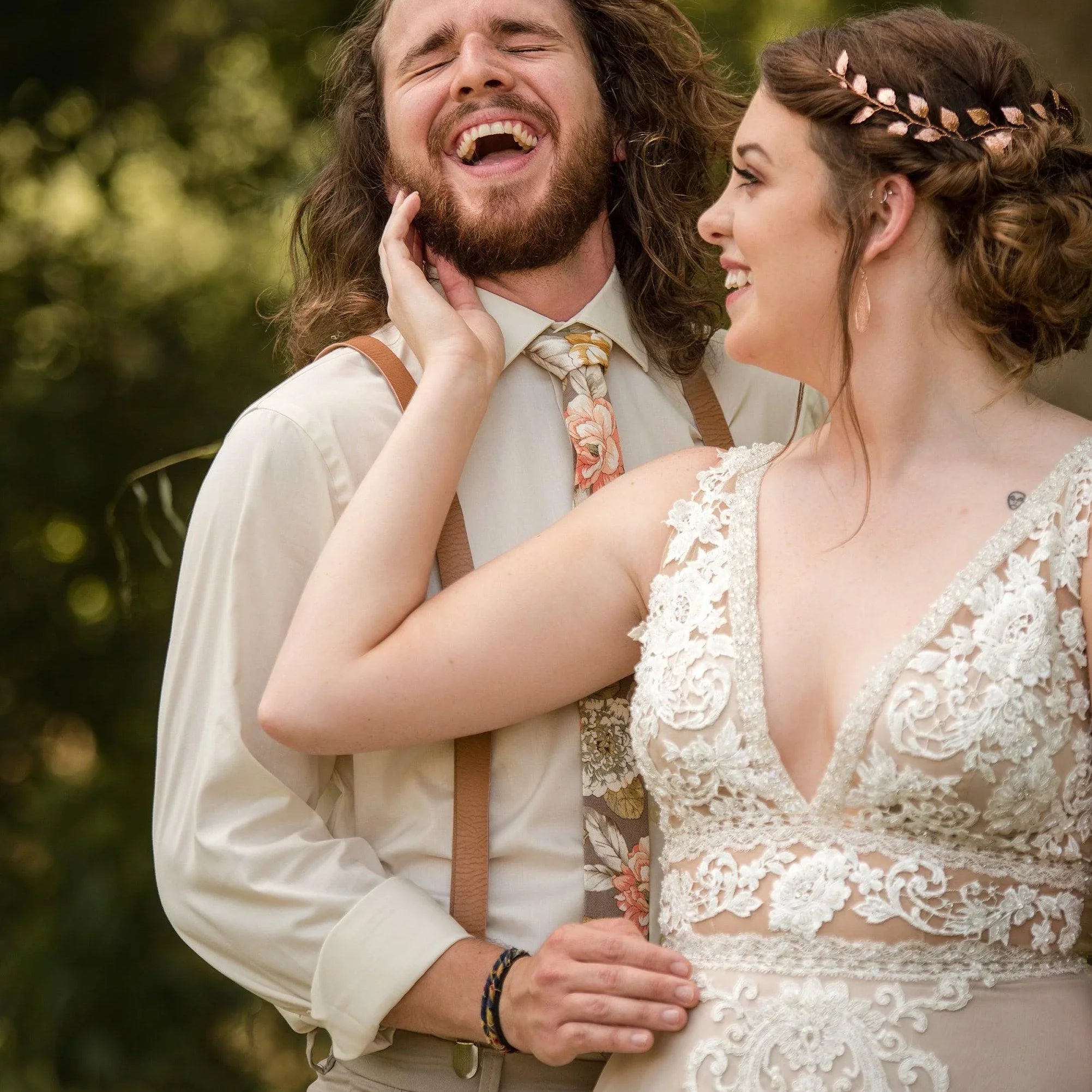 Joyful wedding couple outdoors with the groom in tan suspenders and a champagne floral tie and the bride in a lace wedding dress with a leaf headpiece