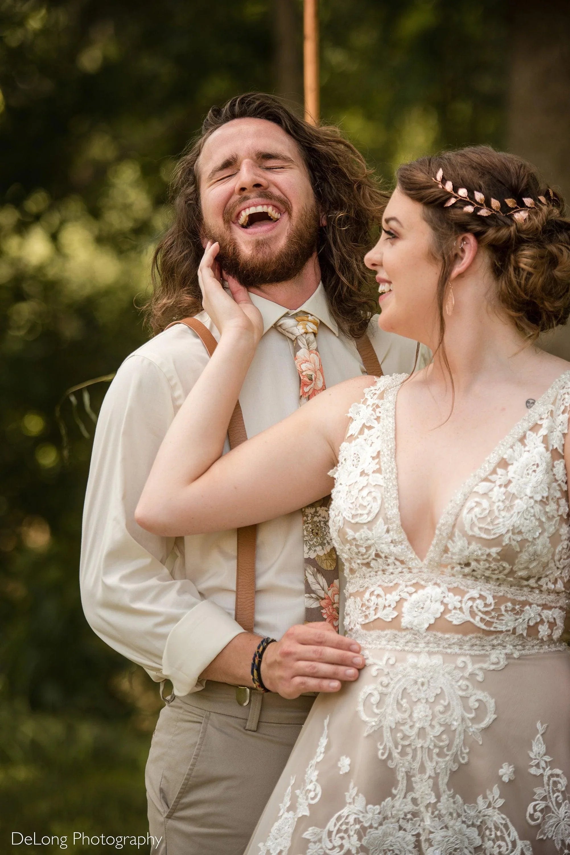 Joyful wedding couple outdoors with the groom in tan suspenders and a champagne floral tie and the bride in a lace wedding dress with a leaf headpiece