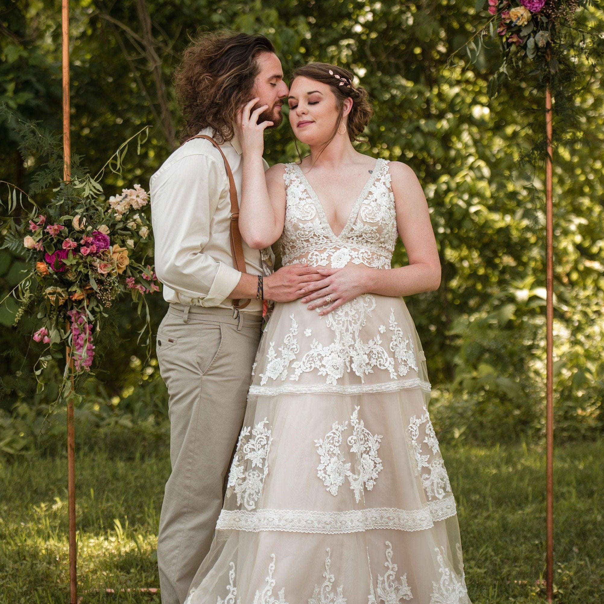 Couple in wedding attire embrace beneath a copper floral arch outdoors