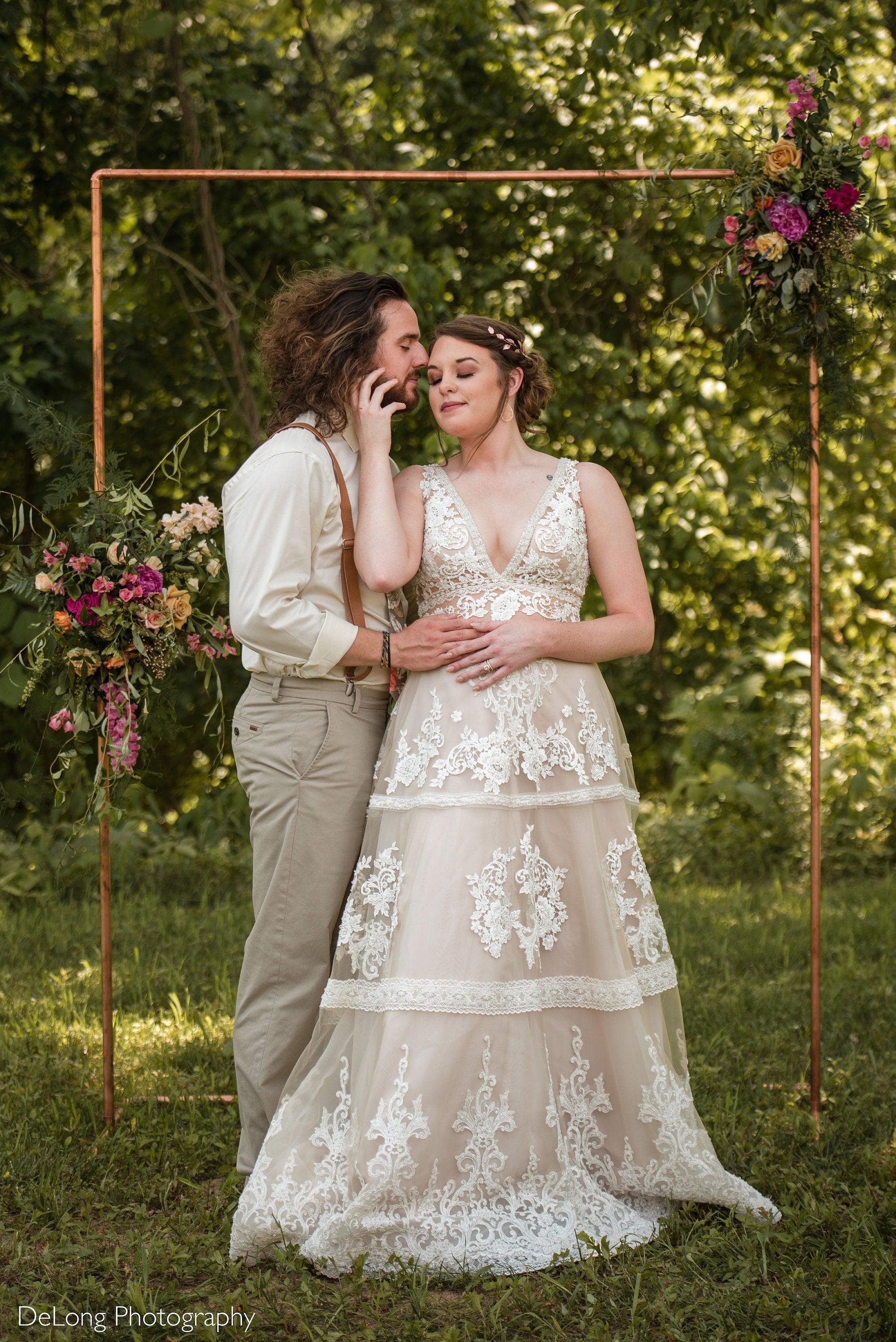 Couple in wedding attire embrace beneath a copper floral arch outdoors