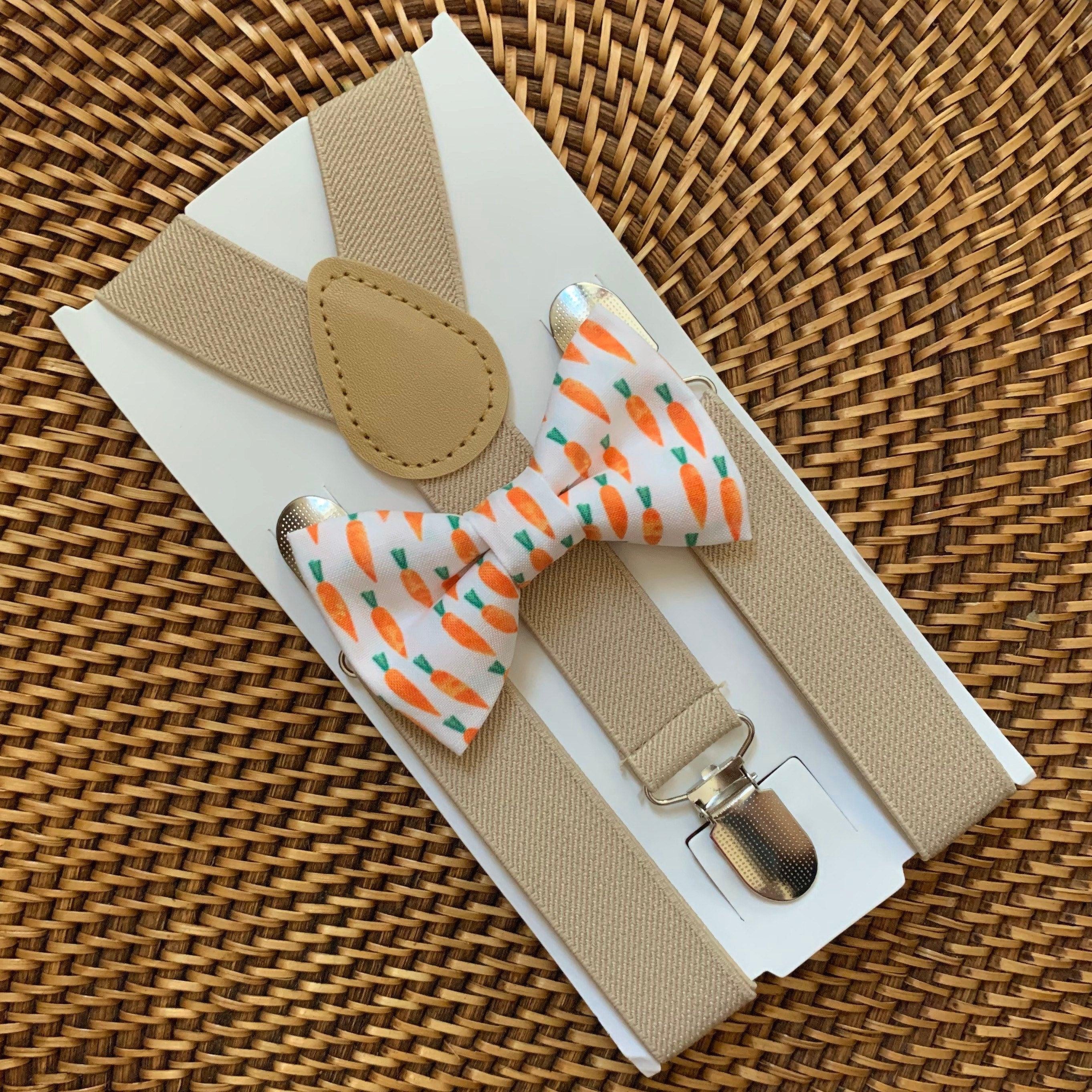 Close up of tan suspenders and carrot print bow tie on a white card against a woven basket background