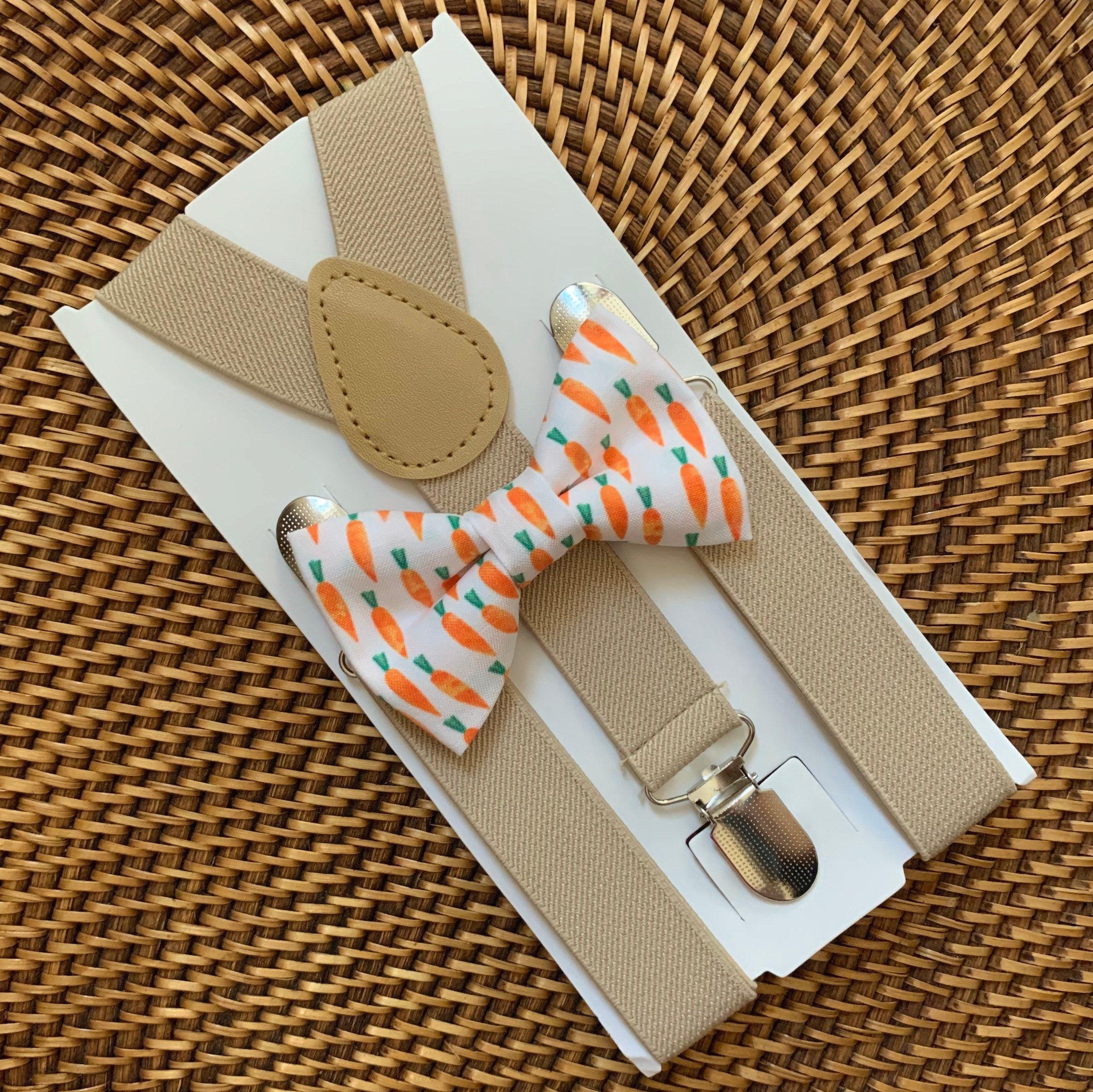 Close up of tan suspenders and carrot print bow tie on a white card against a woven basket background