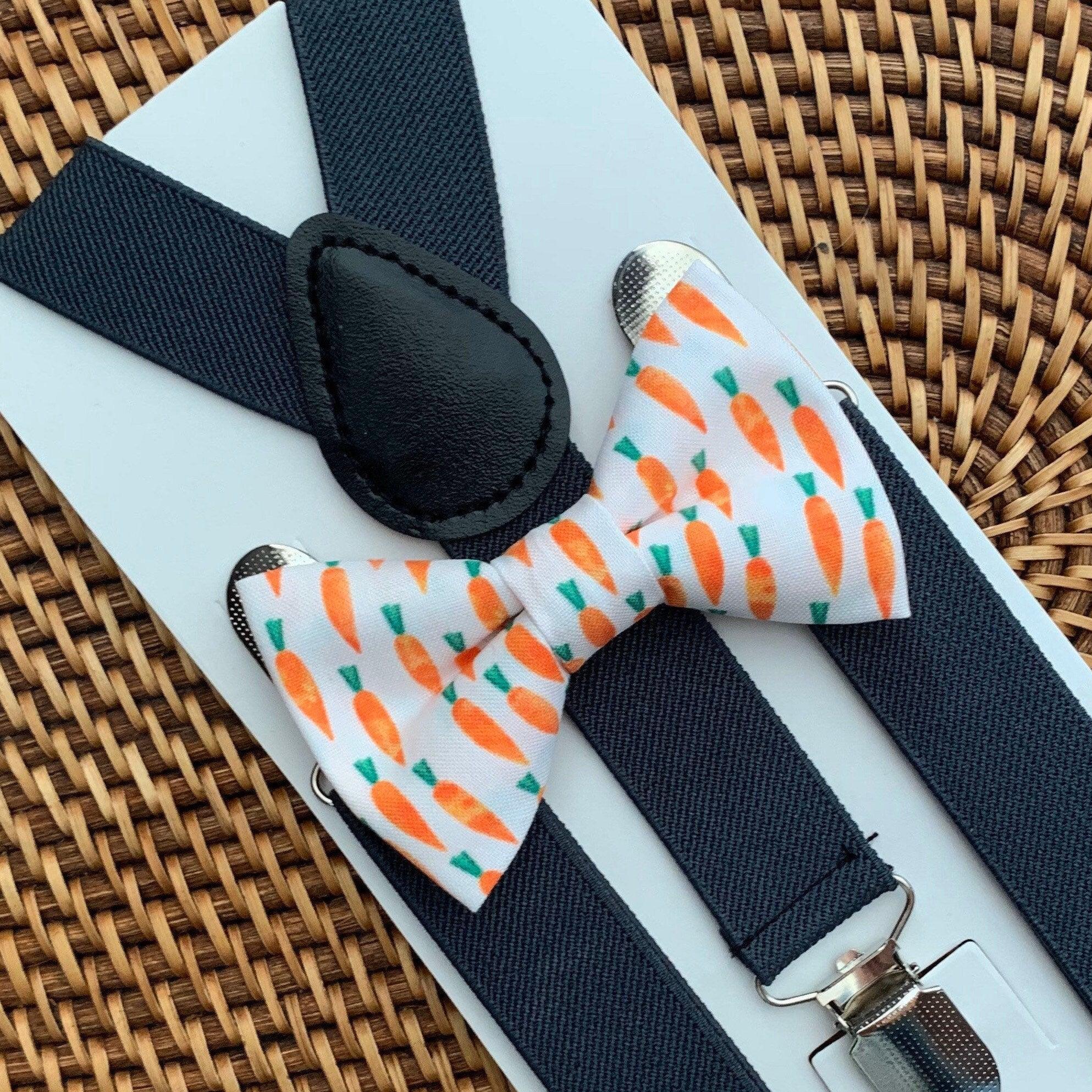 Close up of dark gray suspenders with a stitched leather centerpiece beside a carrot print bow tie on a pale blue display card