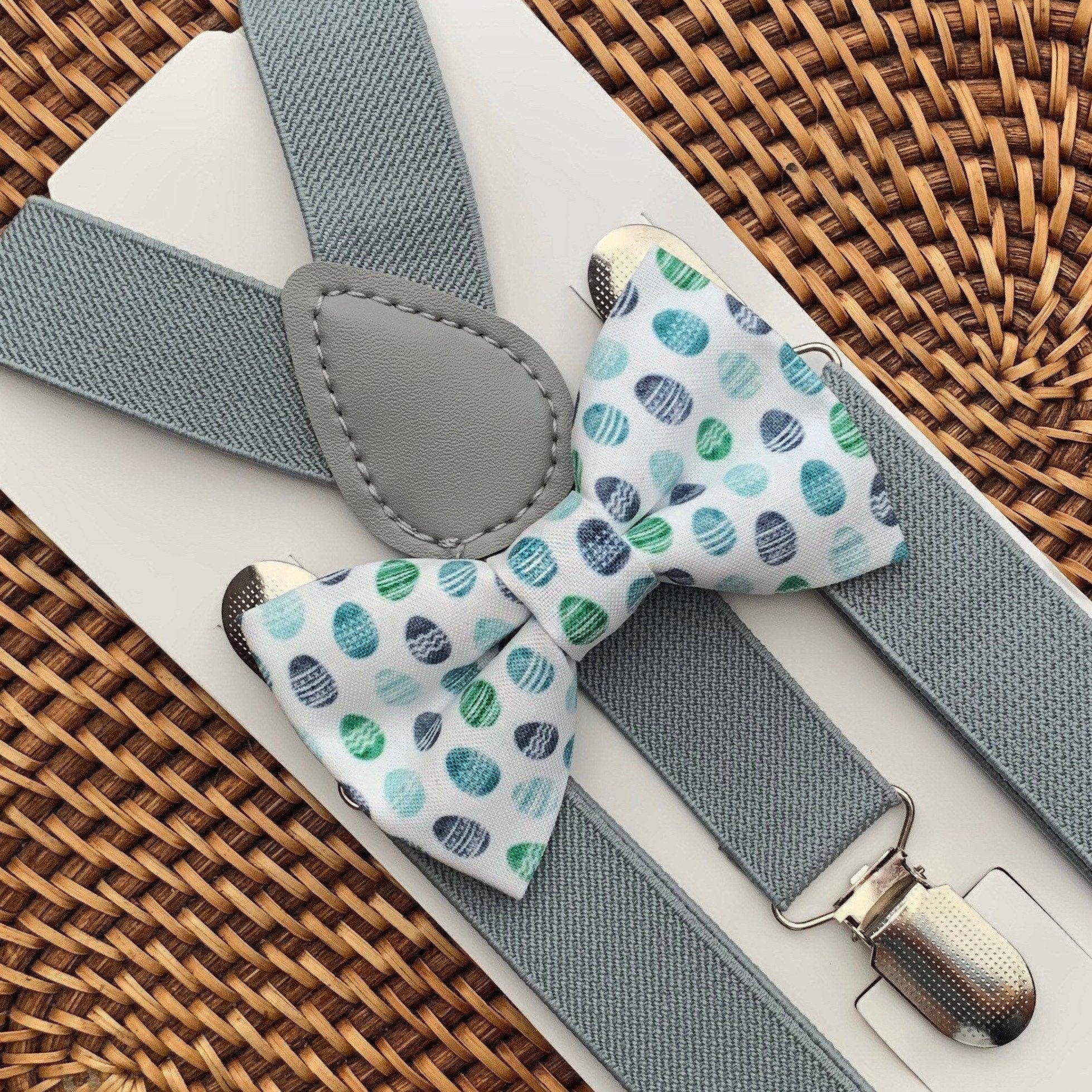 Light gray textured suspenders with a pastel Easter egg patterned bow tie laid on a white surface with a woven basket background.