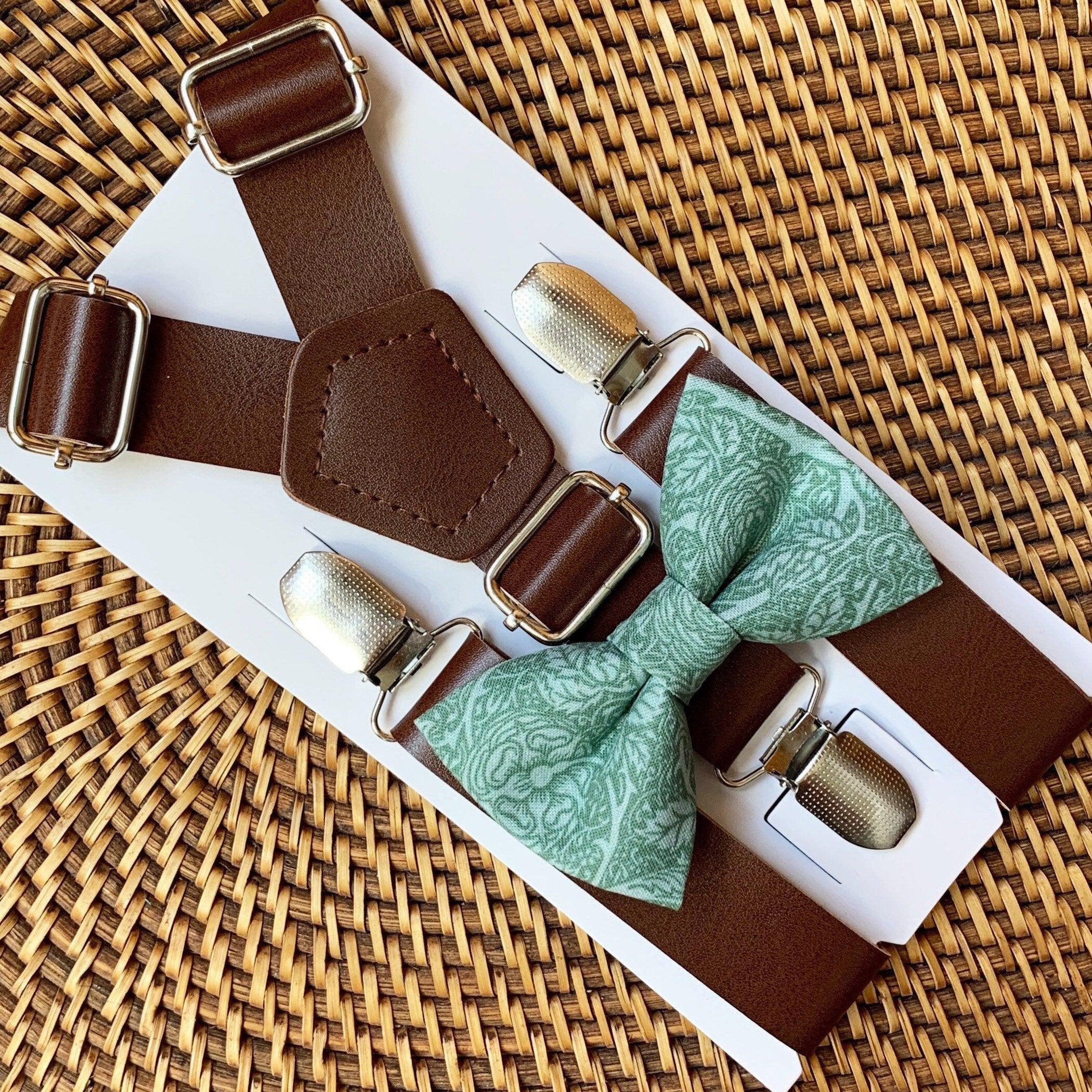 Brown suspenders with silver clips and a green bow tie on a white box against a woven background.