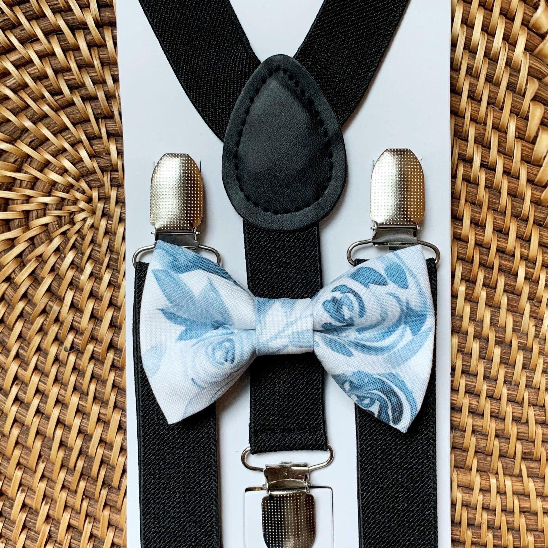A children's suspenders and bow tie set displayed on a white backing card. The suspenders are black with silver metal clips, and the matching bow tie has a light blue and white floral rose pattern.