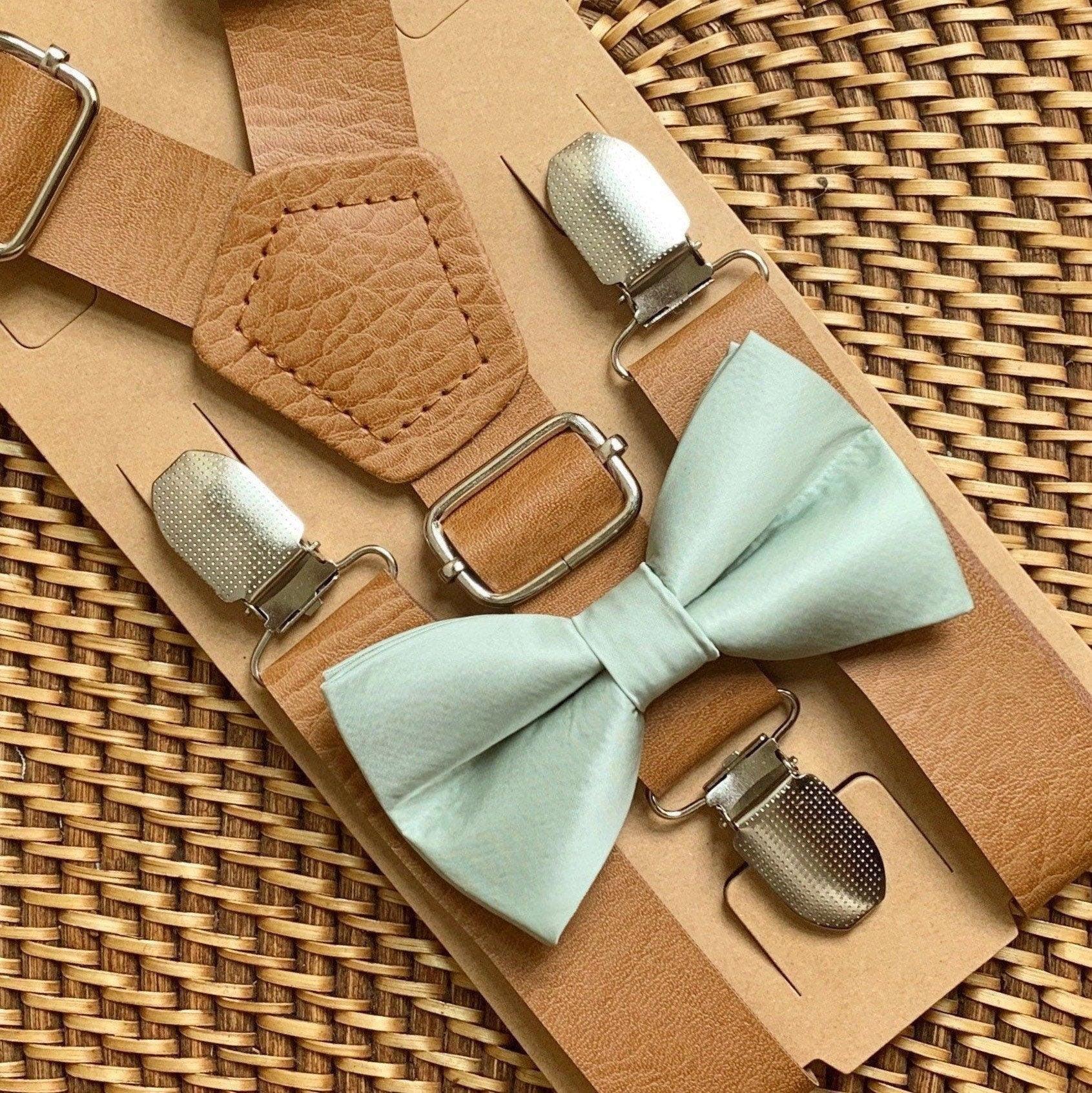 Sage green bow tie with tan vegan leather suspenders laid on a textured brown surface