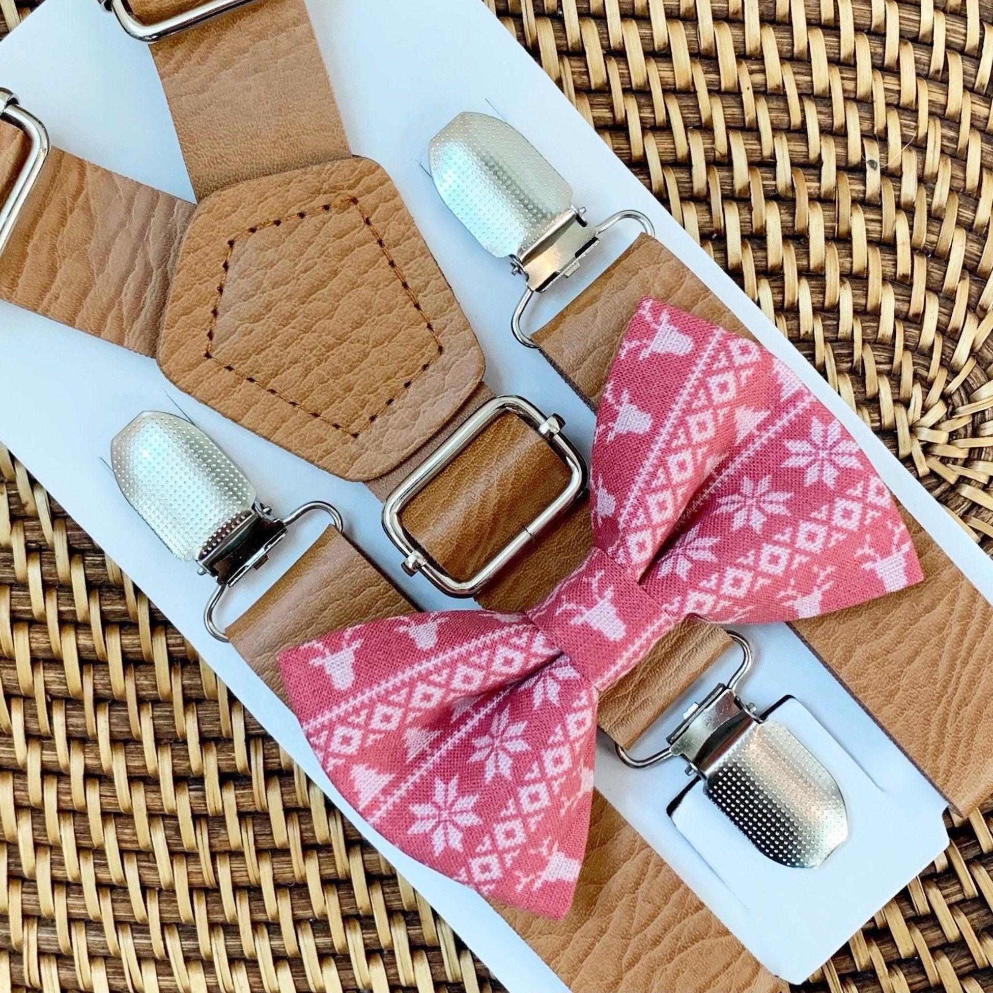 Tan vegan leather suspenders with silver buckles and a pink snowflake bow tie on a white display