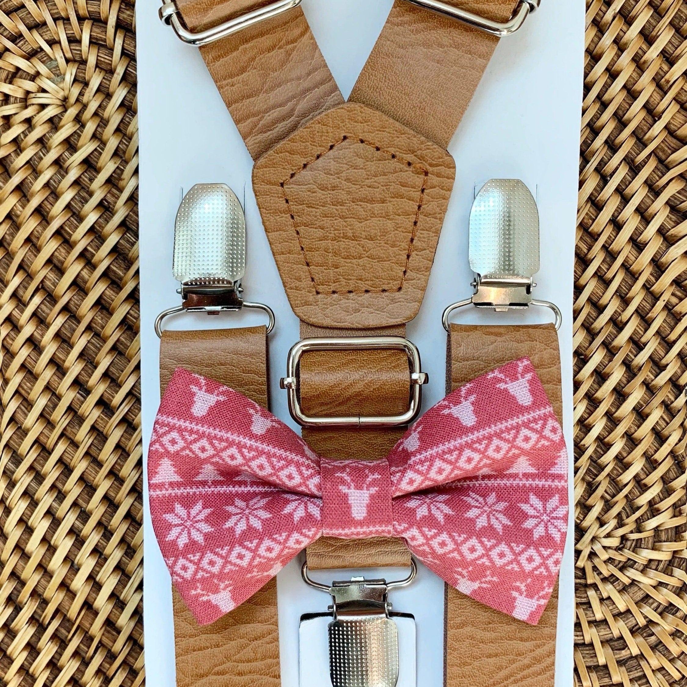 Tan vegan leather suspenders with large metal buckles arranged on a white board with a pink patterned bow tie