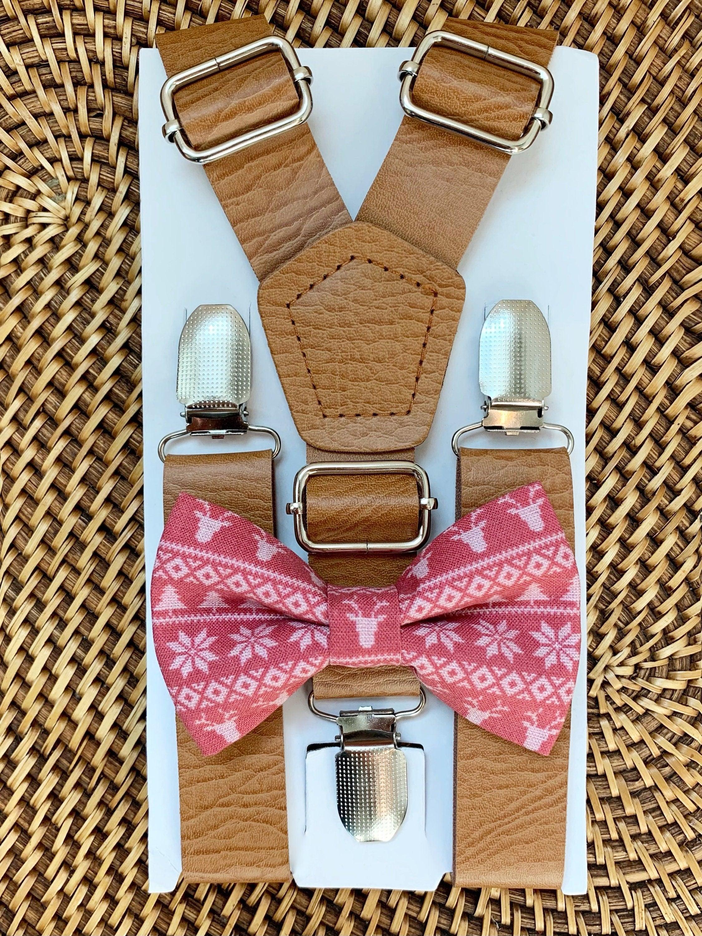 Tan vegan leather suspenders with large metal buckles arranged on a white board with a pink patterned bow tie