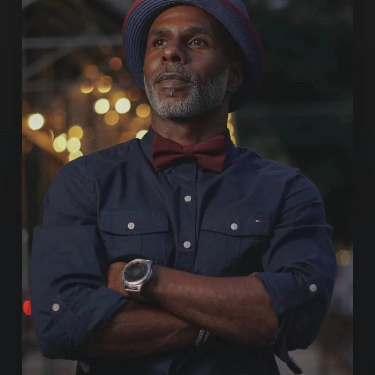 Portrait of a man in a navy shirt wearing a burgundy bow tie with crossed arms and warm bokeh lights behind