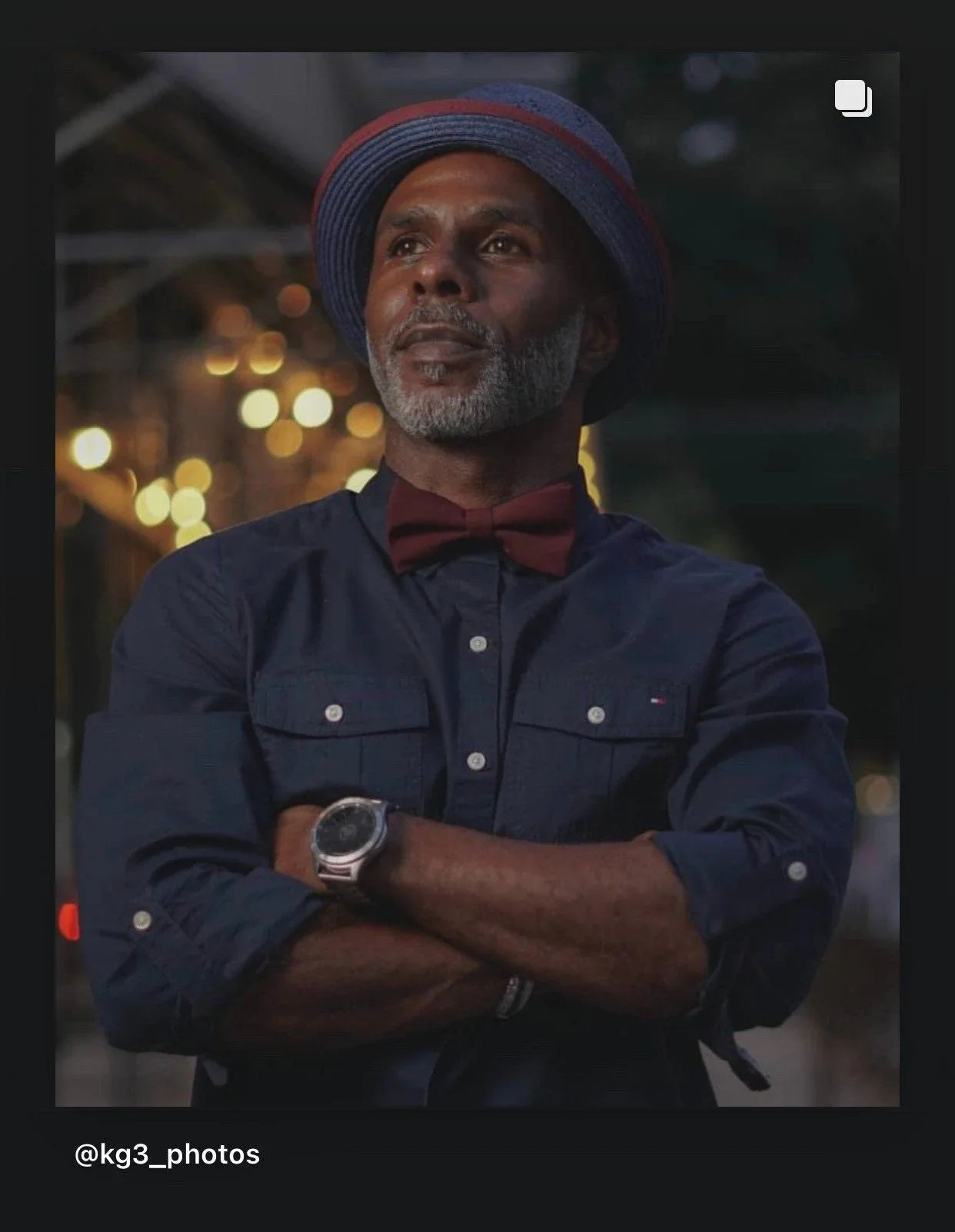 Portrait of a man in a navy shirt wearing a burgundy bow tie with crossed arms and warm bokeh lights behind