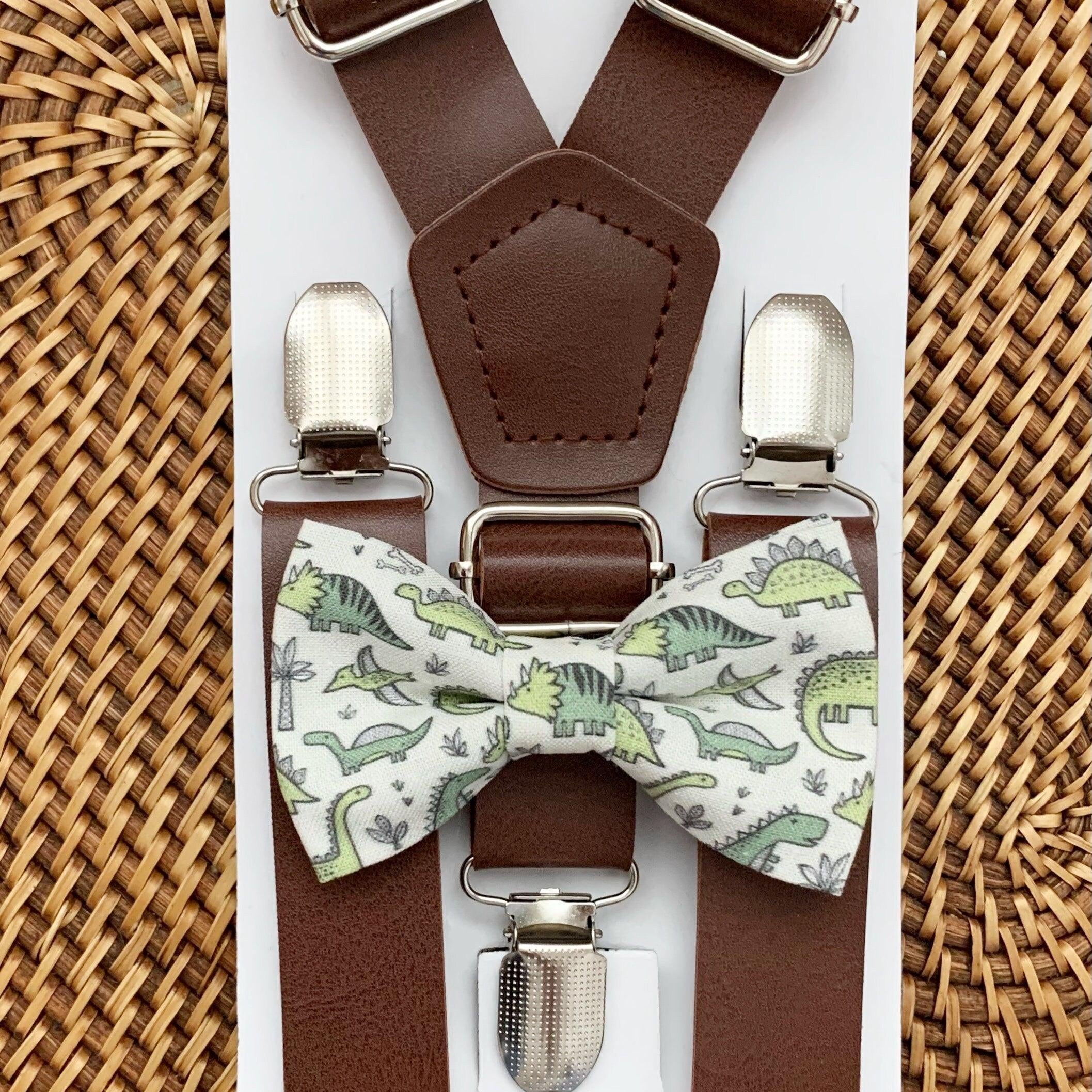 Dark brown vegan suspenders with silver buckles and a green dinosaur patterned bow tie on a white display card