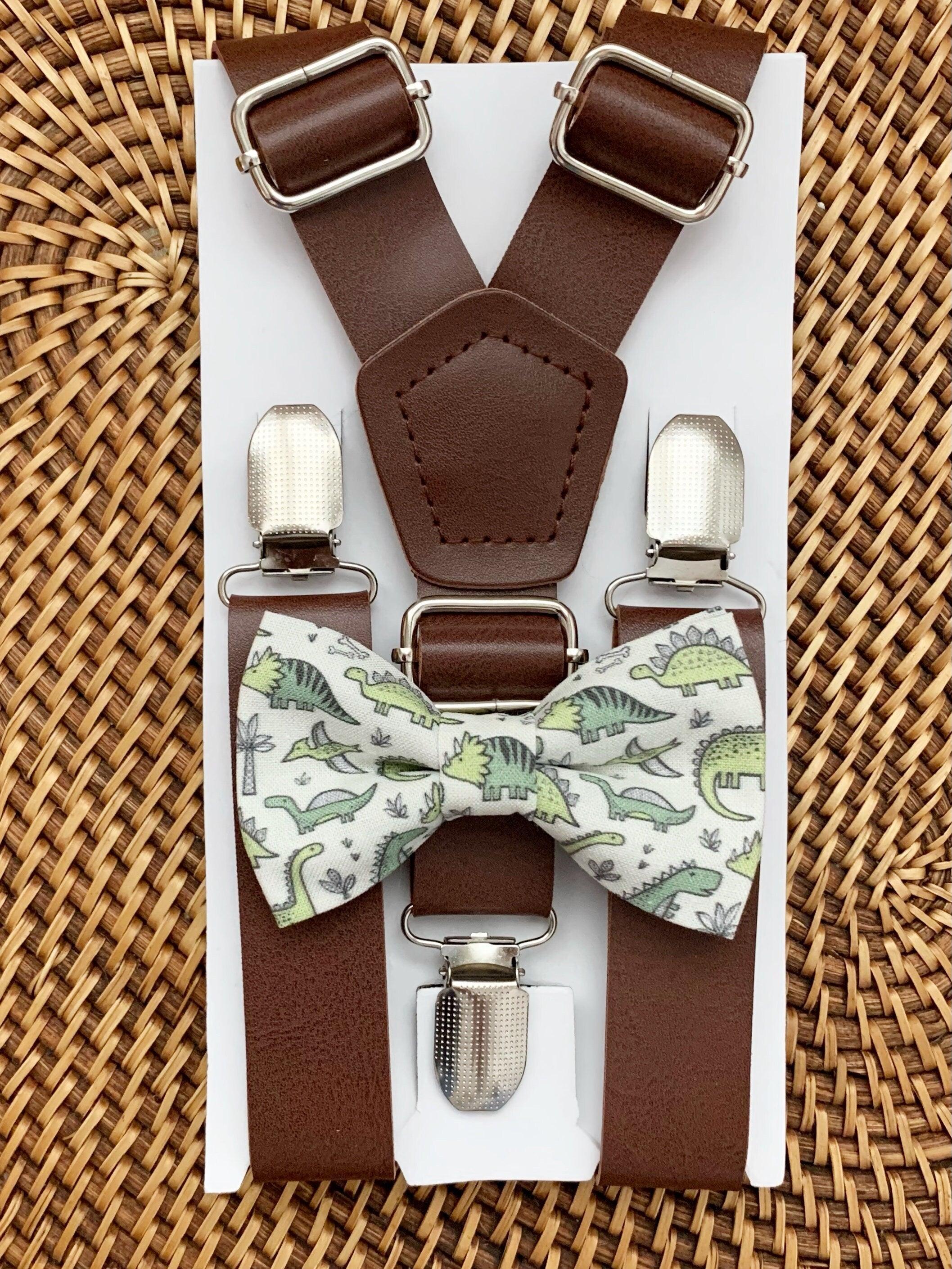 Dark brown vegan suspenders with silver buckles and a green dinosaur patterned bow tie on a white display card