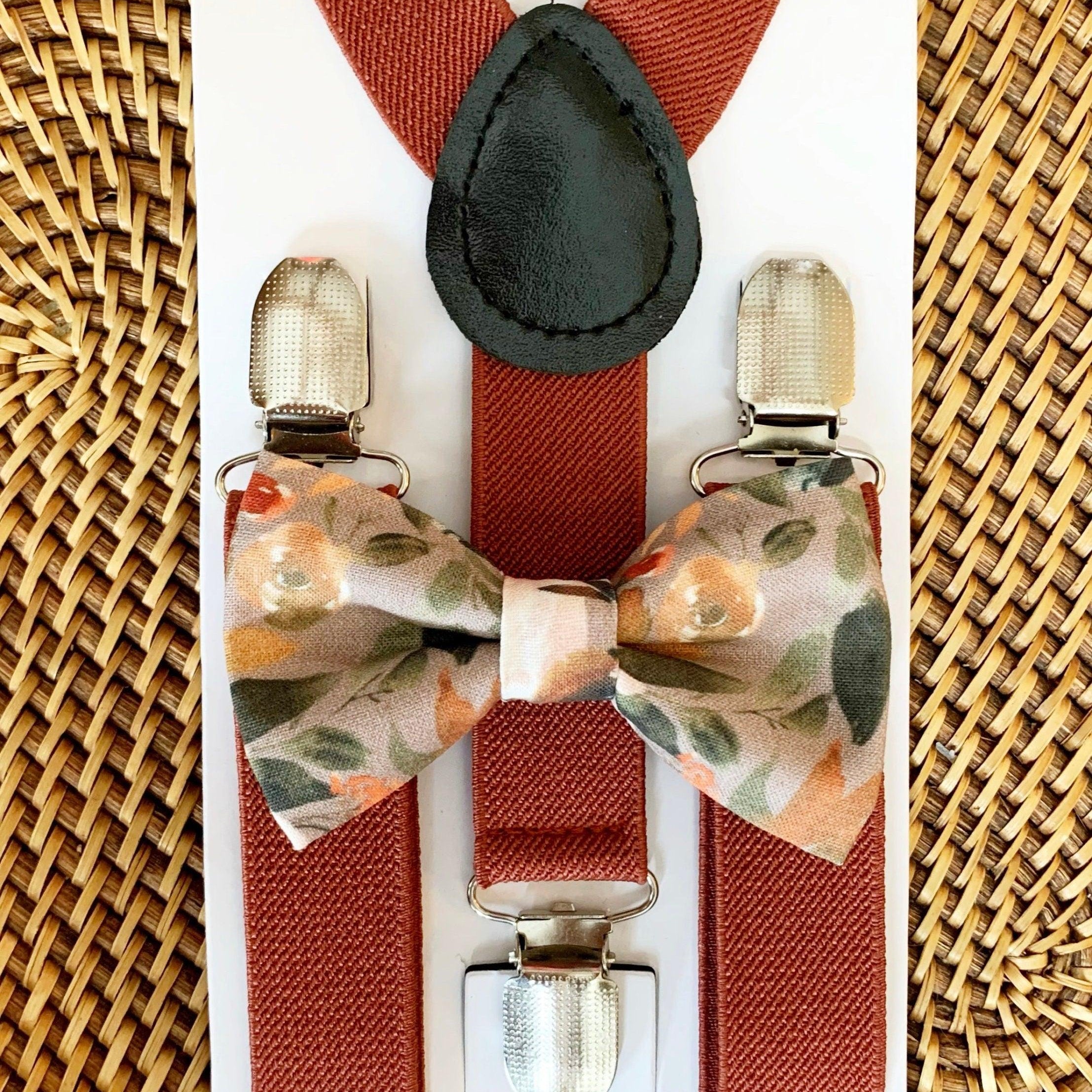 A set of terracotta colored elastic suspenders with a pre-ties autumn floral bow tie displayed on a wooden surface.
