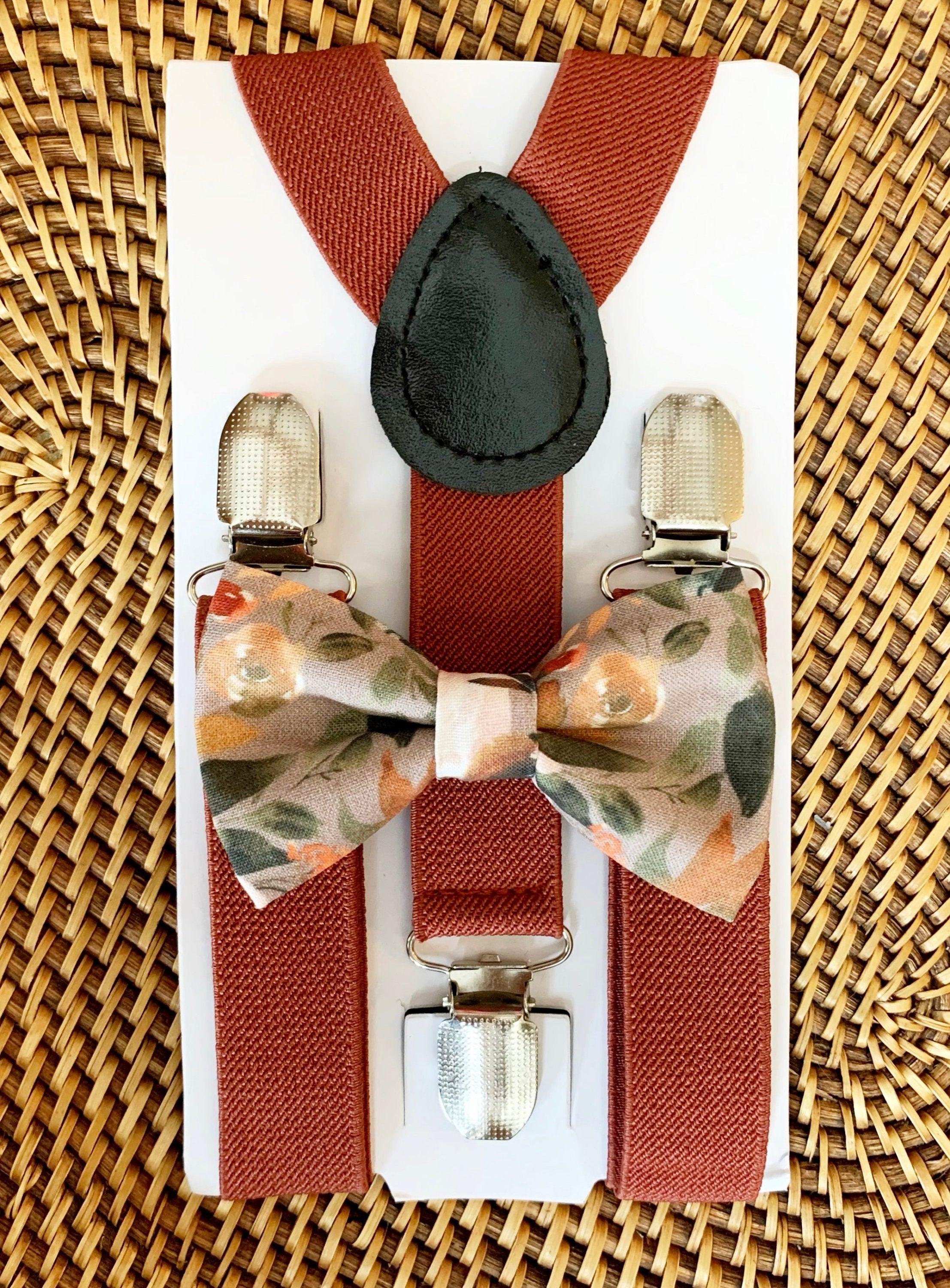 A set of terracotta colored elastic suspenders with a pre-ties autumn floral bow tie displayed on a wooden surface.