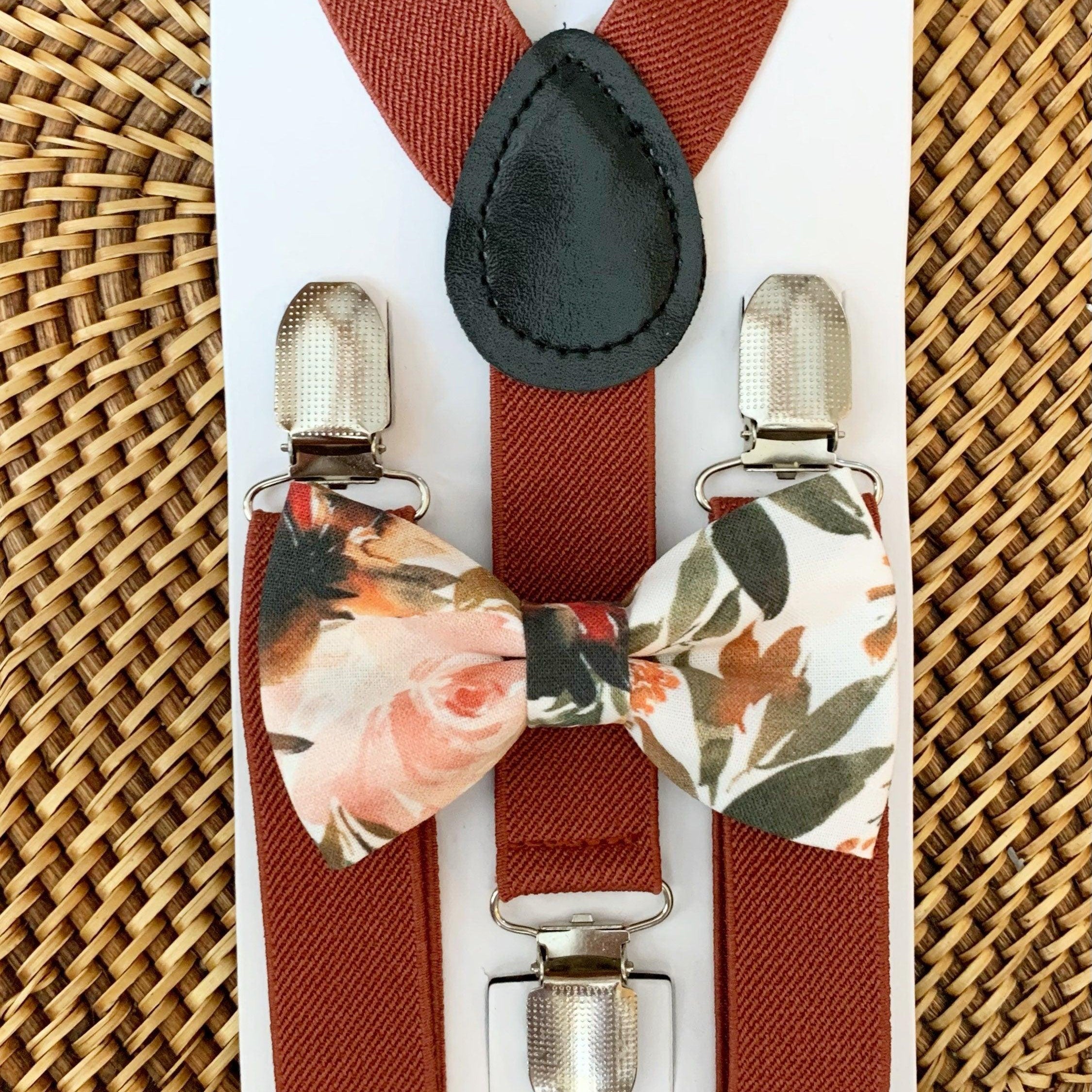 A floral bow tie with ivory, orange, and green colors on a white background, paired with cinnamon rust colored suspenders with metal clips, displayed on a woven surface.