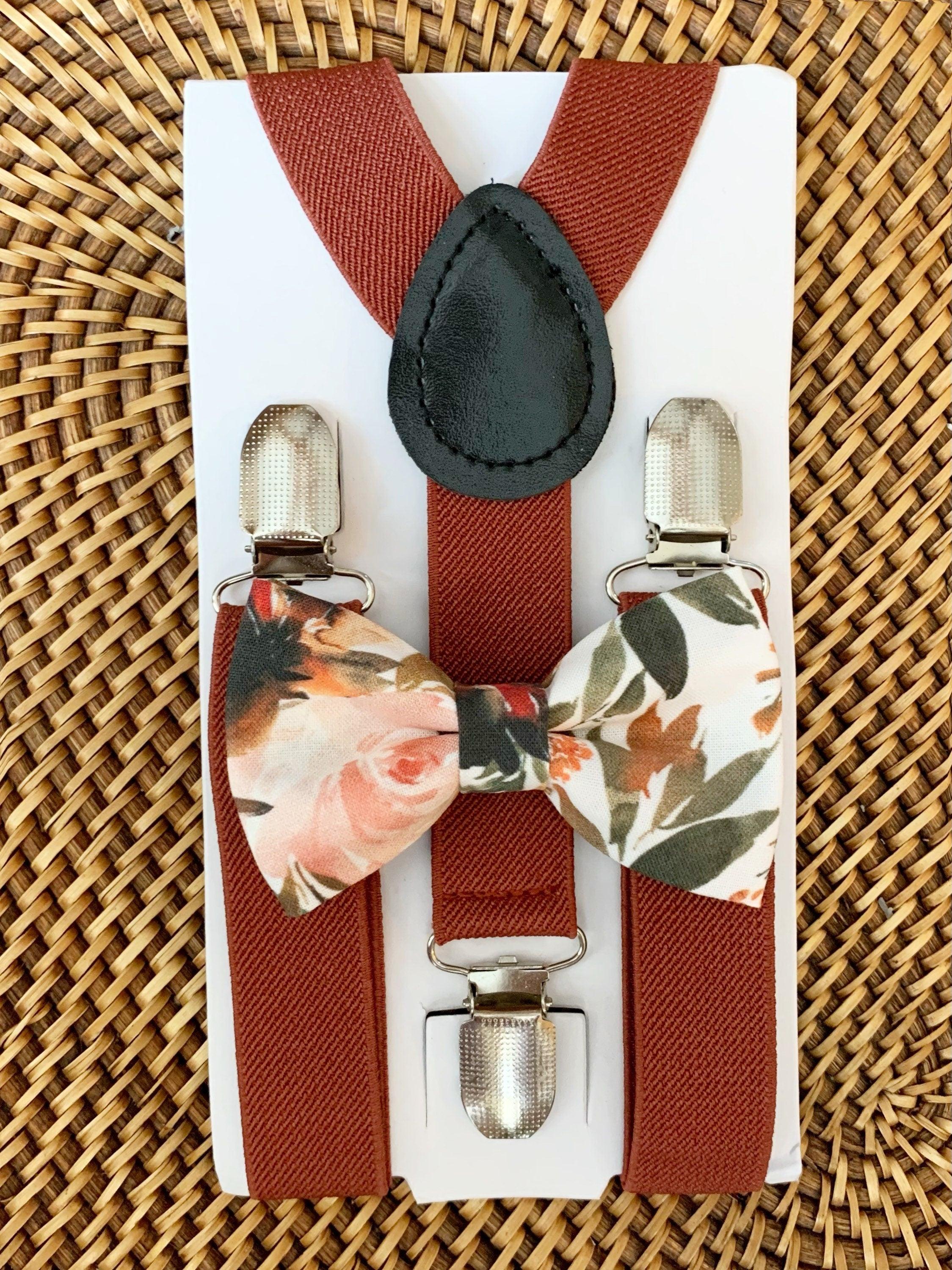 A floral bow tie with ivory, orange, and green colors on a white background, paired with cinnamon rust colored suspenders with metal clips, displayed on a woven surface.
