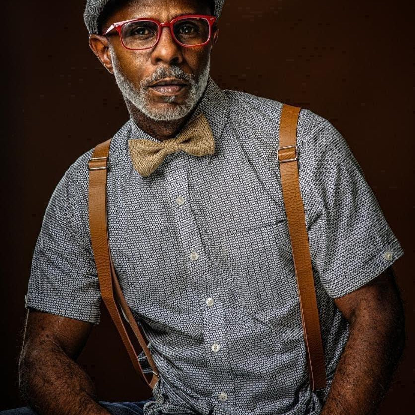 A handsome man in red glasses and a page boy hat wears a country chic burlap bow tie and rustic tan suspenders for men and boys to wear to a cowboy wedding.