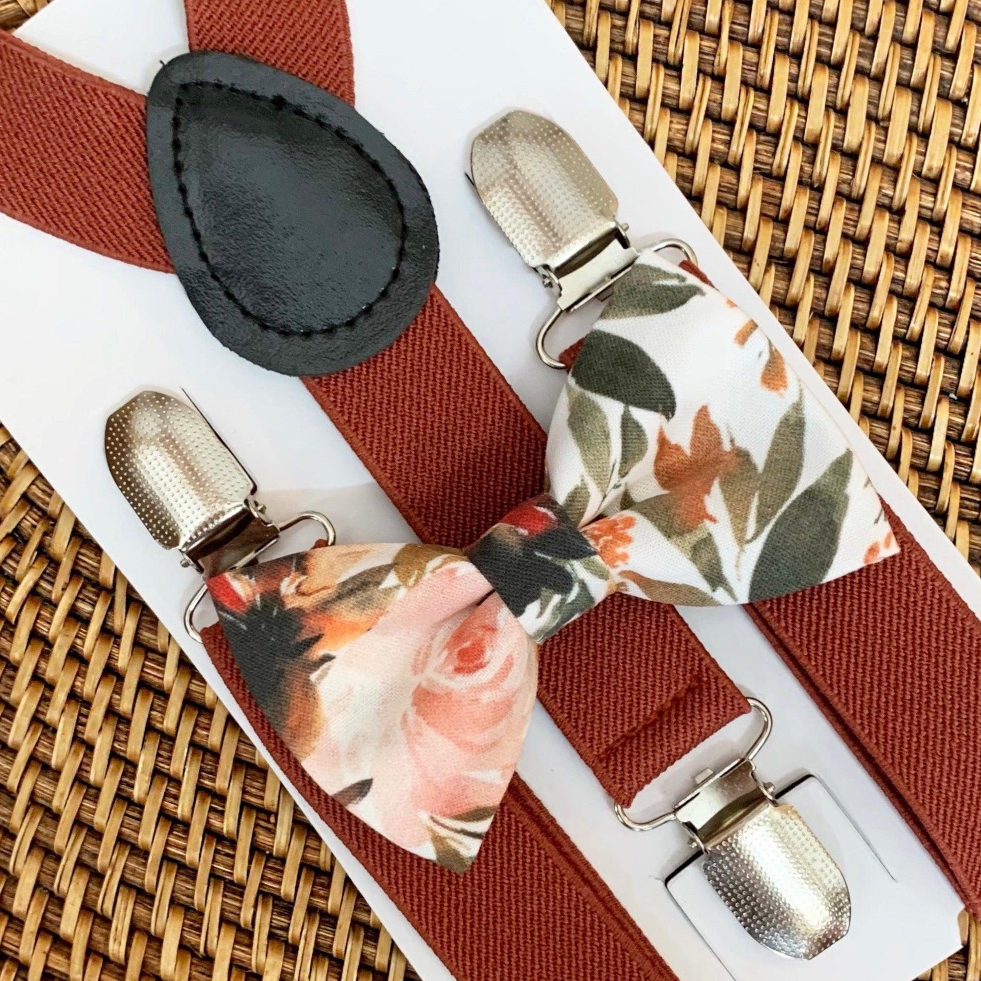 A floral bow tie with ivory, orange, and green colors on a white background, paired with cinnamon rust colored suspenders with metal clips, displayed on a woven surface.