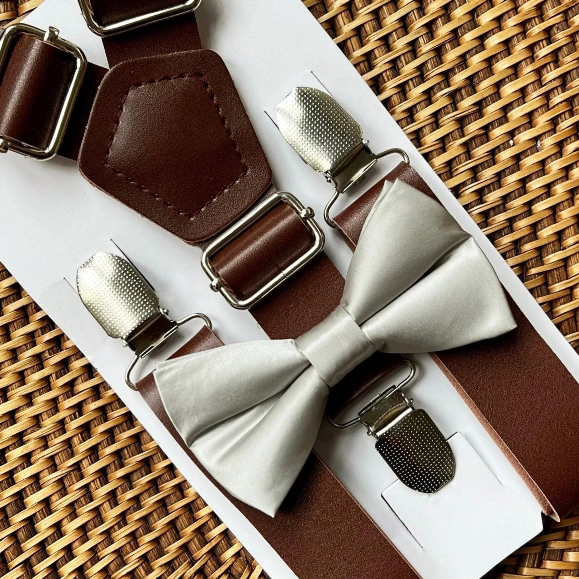 Brown suspenders with silver clips and a silver bow tie on a woven surface
