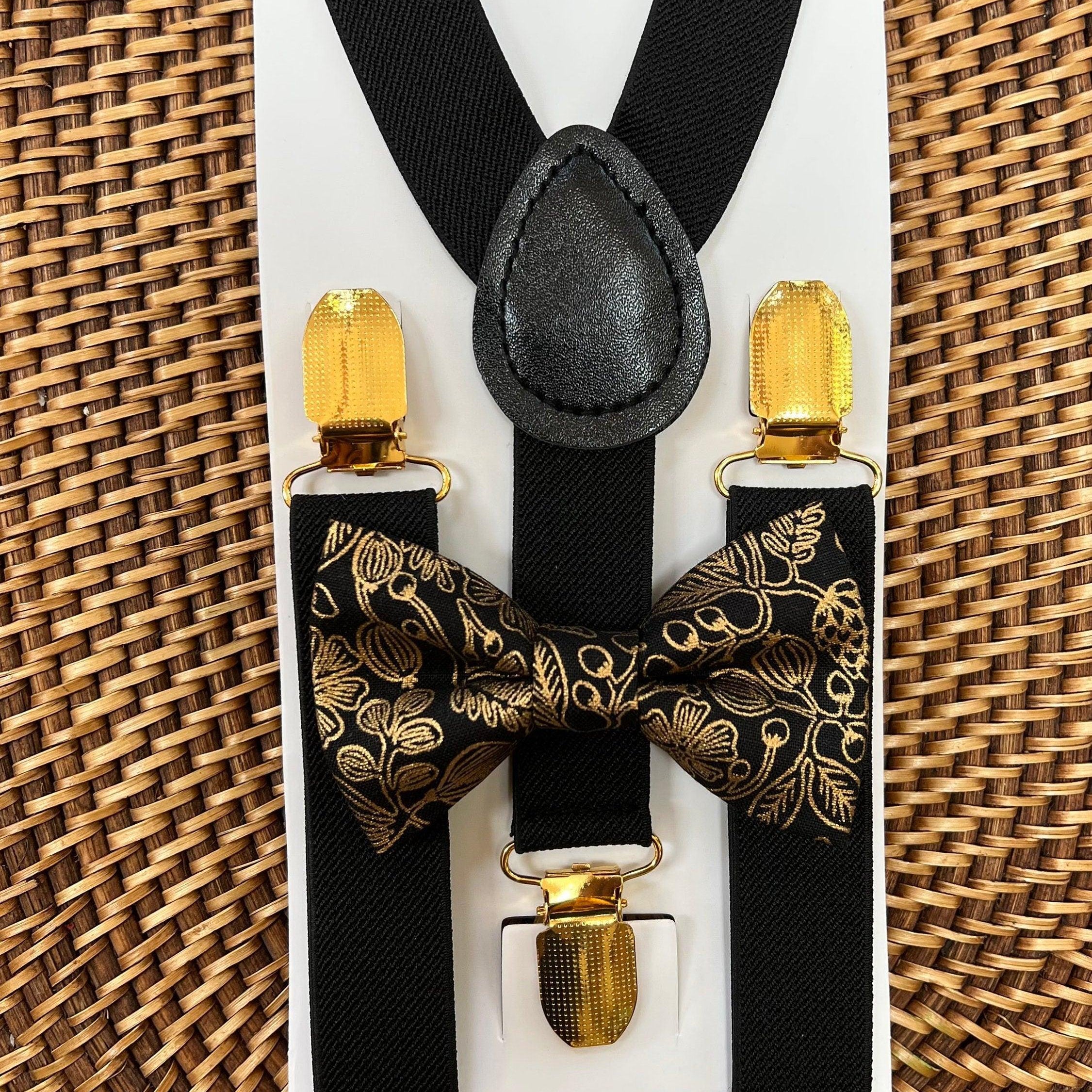 Black and gold bow tie and suspenders set shown on a white card with gold clip ends and a leather knot centerpiece