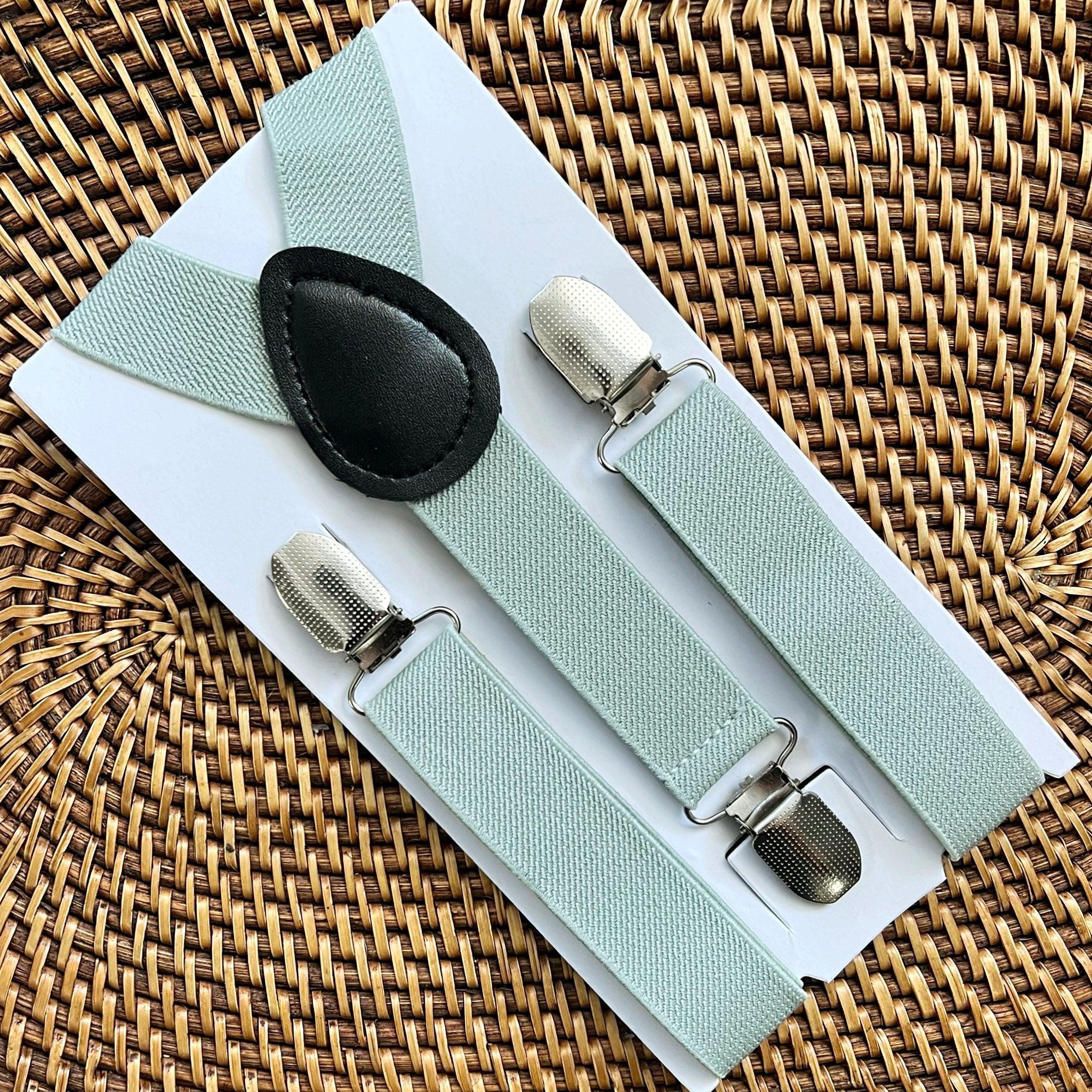 Close up of sage green fabric suspenders with metal clips on a pale display card over a woven wicker background